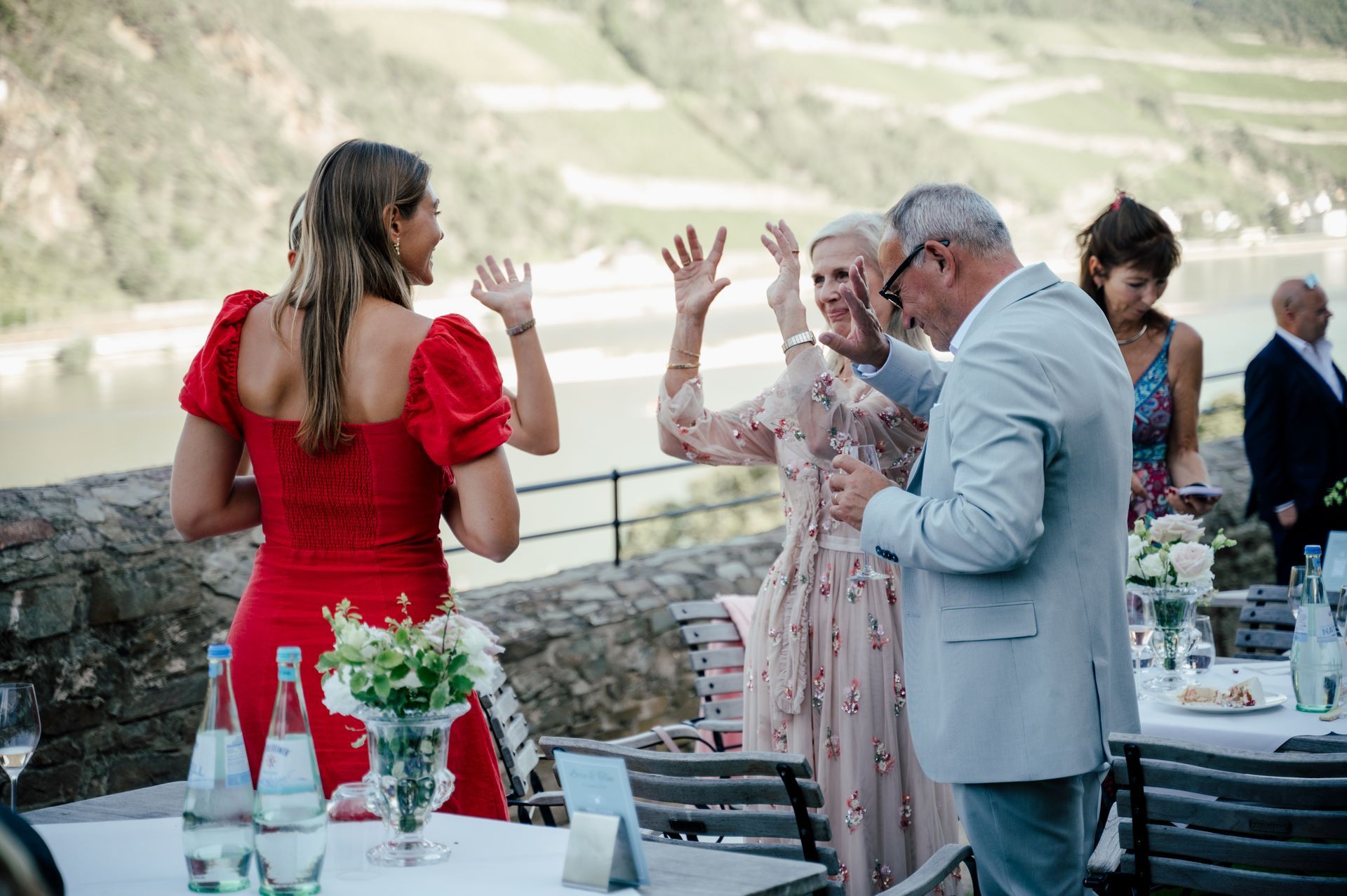 Four people at a sunny outdoor gathering by a river, with some of the participants waving to each other across the tables.