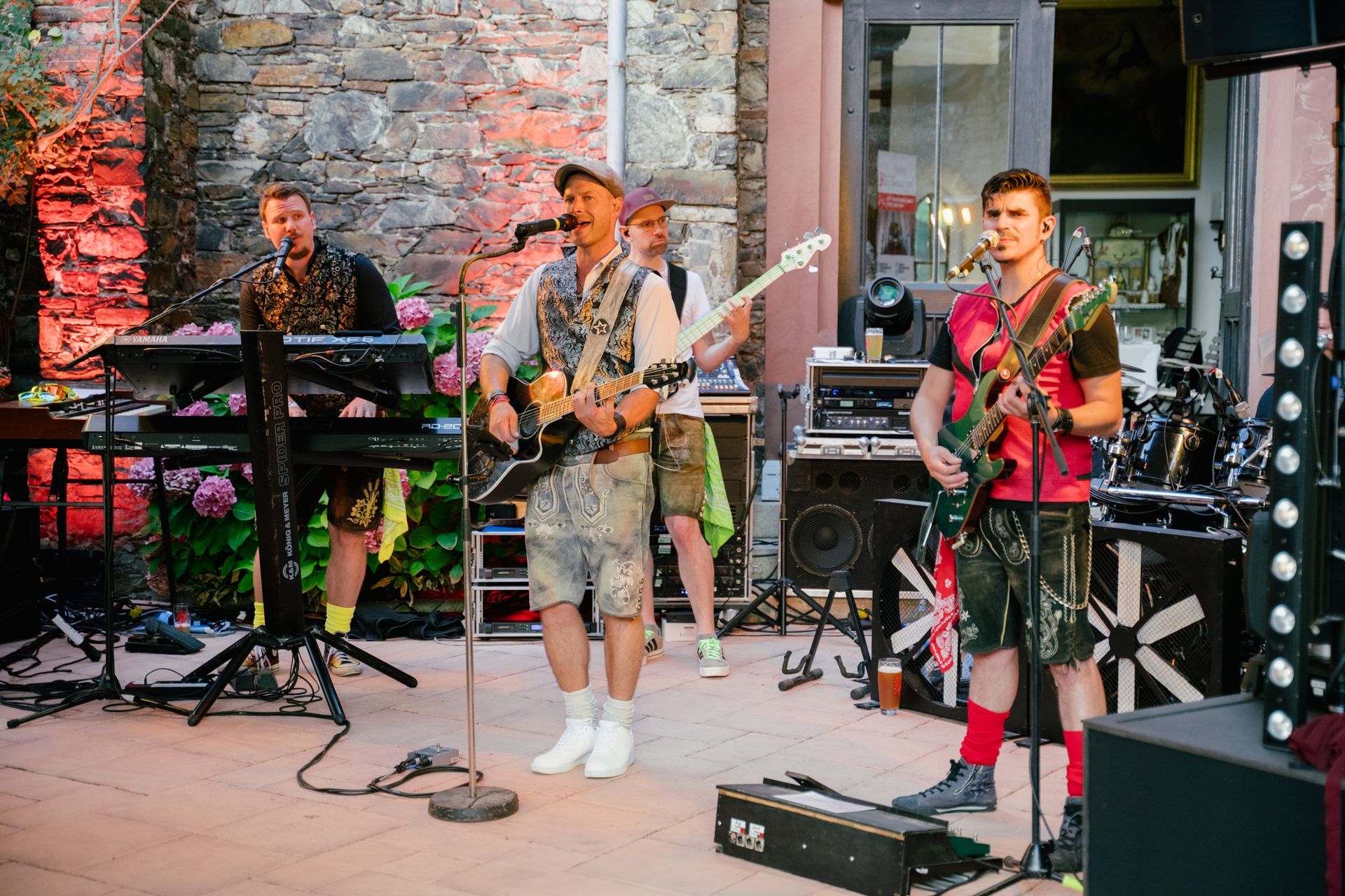 A band is performing live on a terrace with stone walls, playing the keyboard and guitar and singing into microphones.