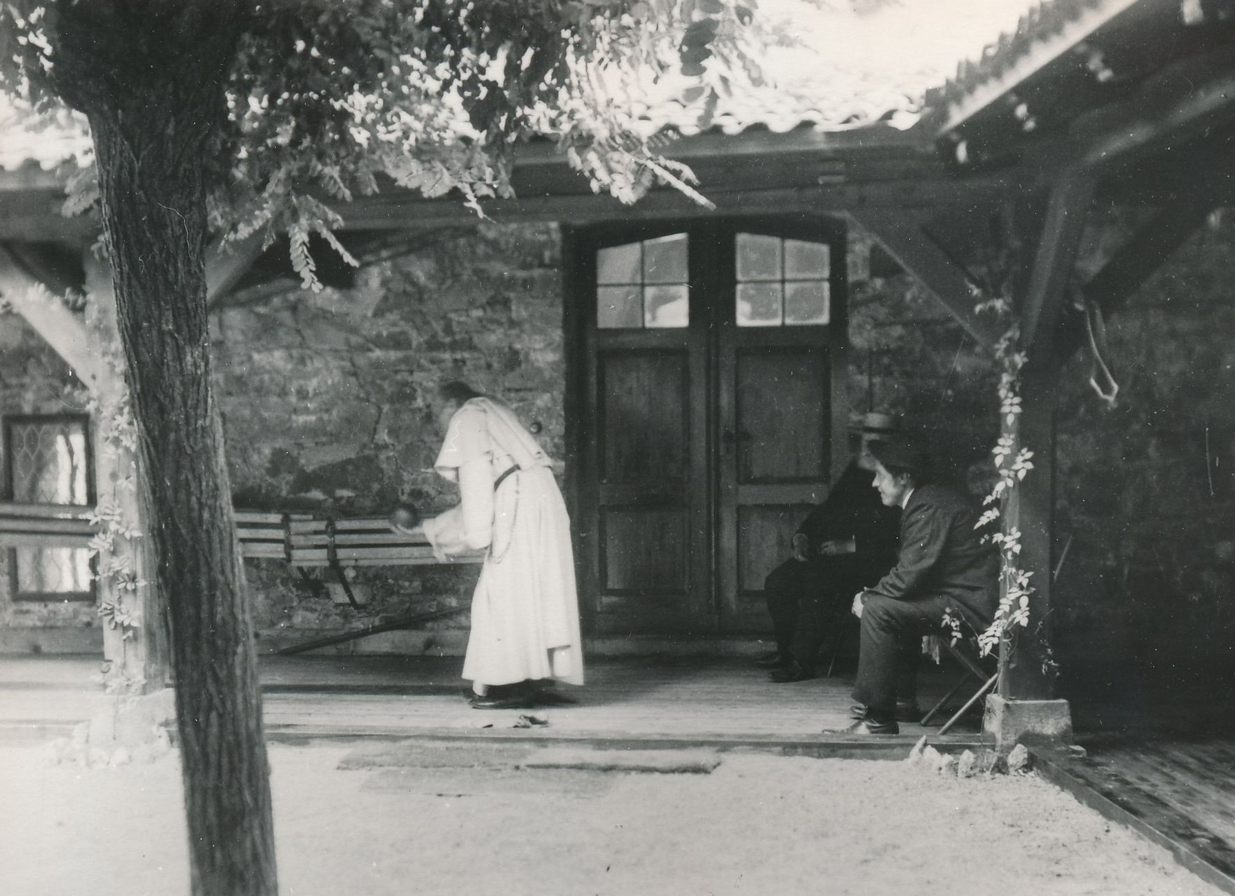 Eine Person in einem langen Gewand steht auf einer steinernen Veranda in der Nähe eines Gebäudeeingangs, während eine andere Person daneben auf einem Holzstuhl sitzt.