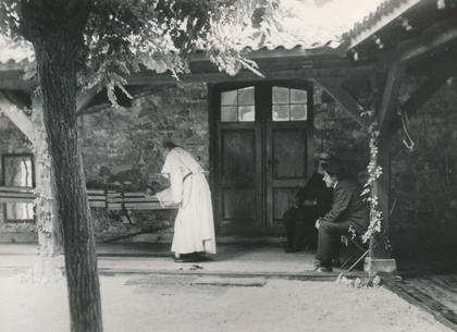 Eine Person in einem langen Gewand steht auf einer steinernen Veranda in der Nähe eines Gebäudeeingangs, während eine andere Person daneben auf einem Holzstuhl sitzt.