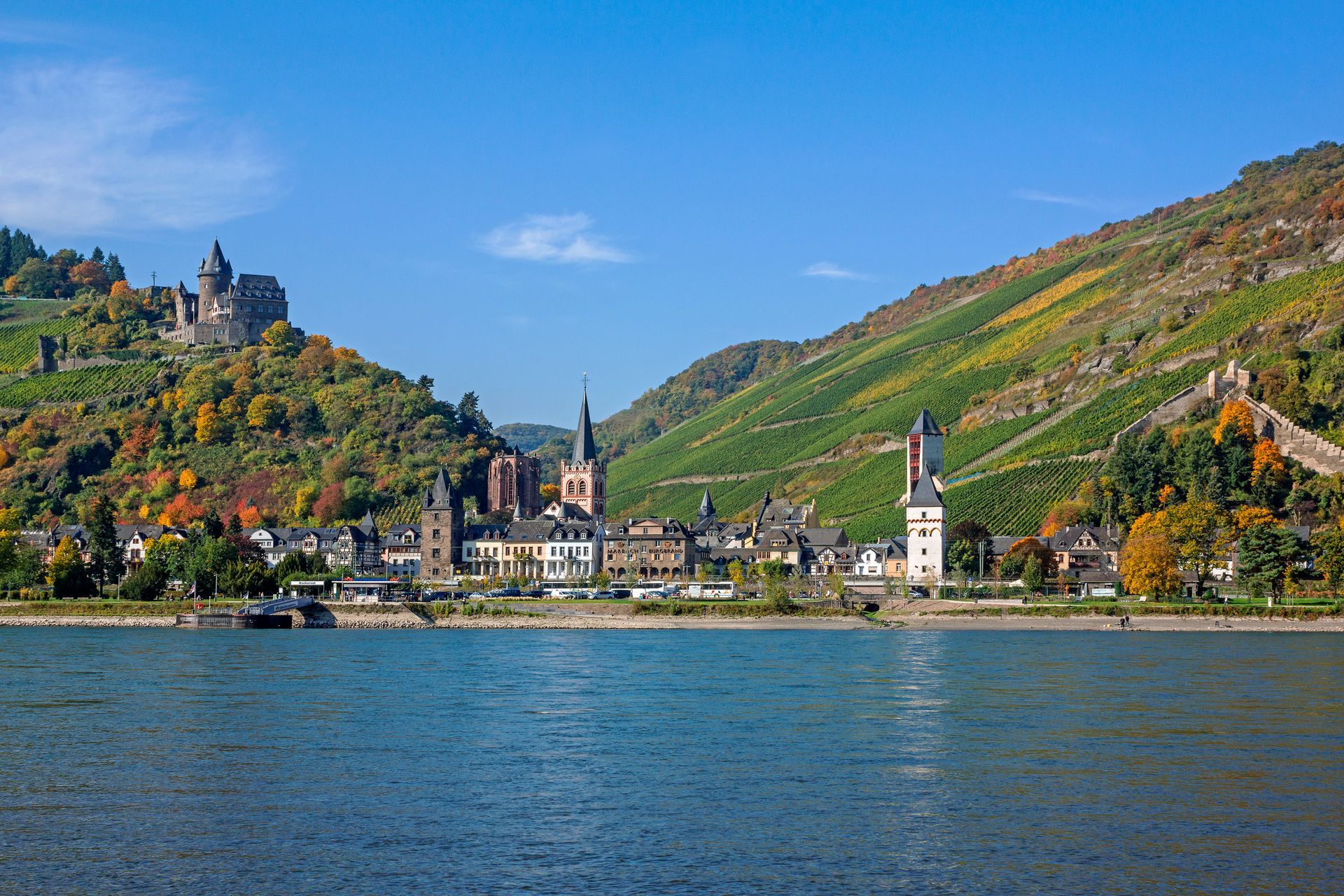 Blick auf die Stadt Bacharach am Rhein mit Schloss Stahleck, üppigen Terrassenweinbergen und blauem Himmel.