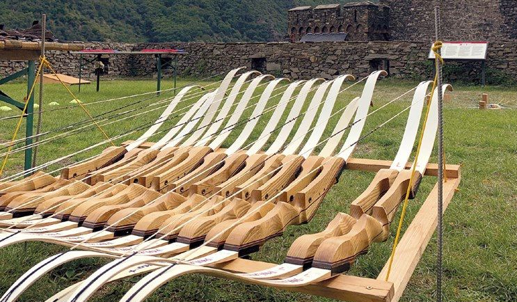 Several wooden recurve bows were lined up on a wooden rack in a grassy open area; a stone wall could be seen in the background.