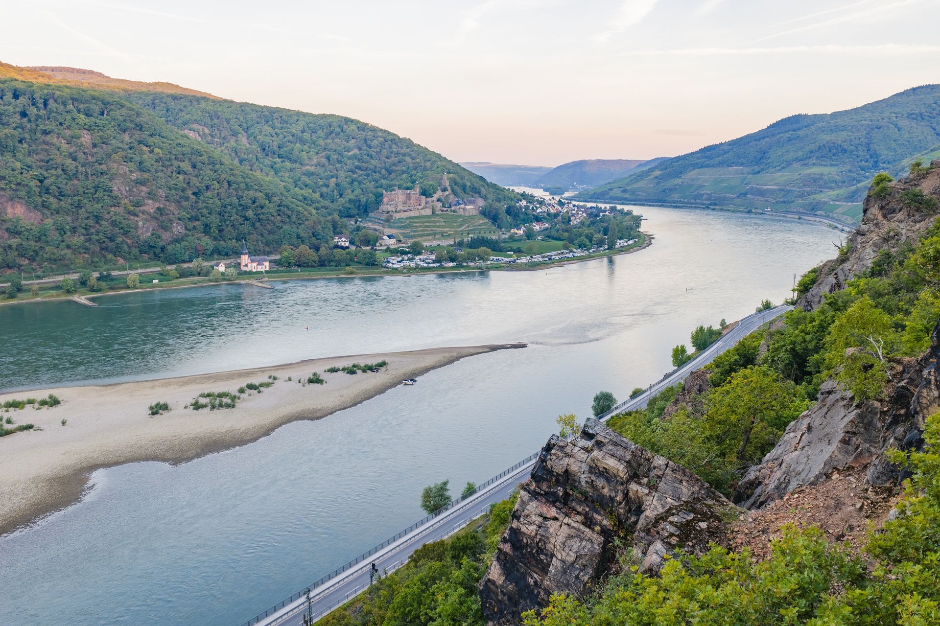 Ein breiter, fließender Fluss schlängelt sich durch ein grünes Tal, im Vordergrund eine Sandbank und entlang des felsigen Ufers eine Straße.