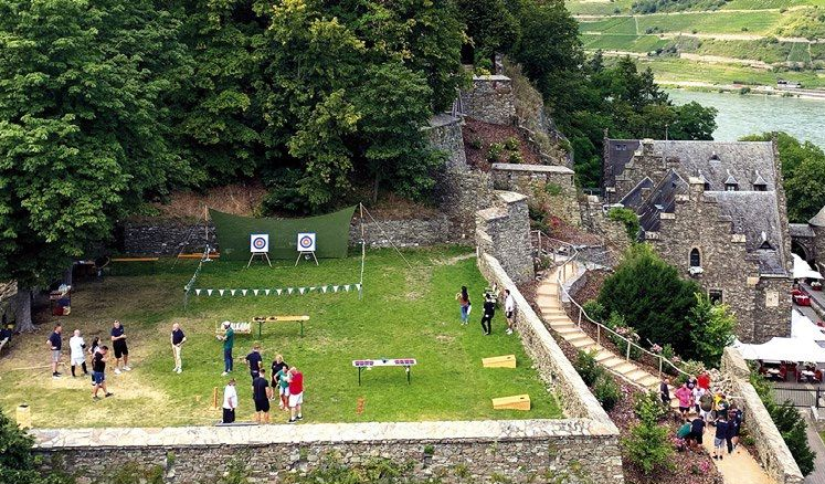 Ein Bogenschießstand im Innenhof einer historischen Steinburg, auf dem sich Menschen auf dem Rasen in der Nähe der Zielscheiben versammelt haben.