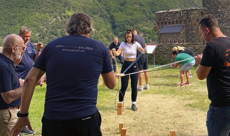A group of people outdoors are pulling on ropes to operate a device and help a person who is balancing on wooden blocks.