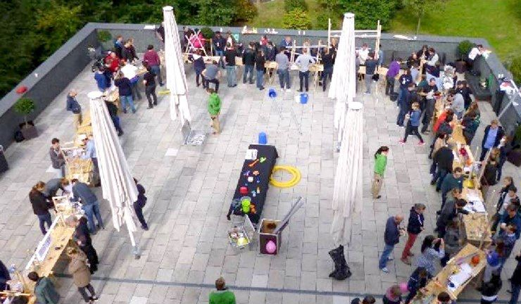 An outdoor gathering on a tiled patio with people sitting around tables, large white umbrellas, and a black play mat.