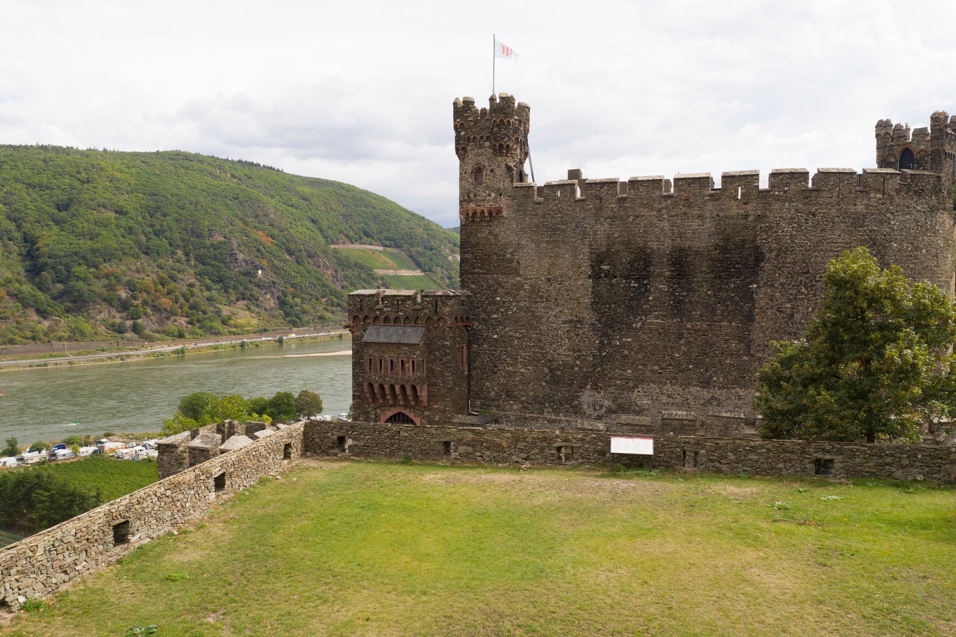 Perched on a grassy hill stands a historic stone castle with a tower, overlooking a wide river valley with green, wooded slopes.