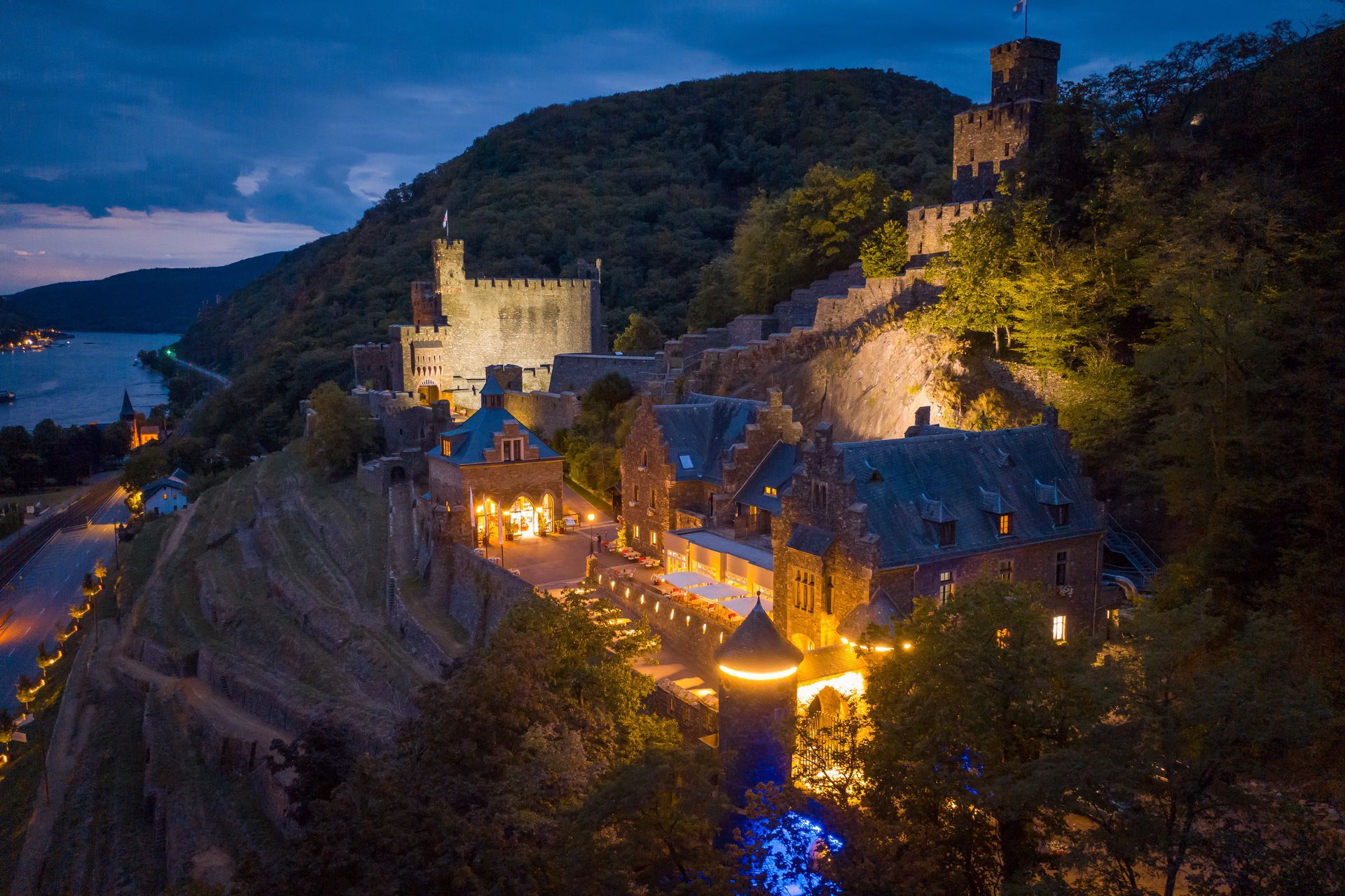Eine beleuchtete Burganlage, die auf einer Felsklippe thront und in der Abenddämmerung einen Fluss überblickt.