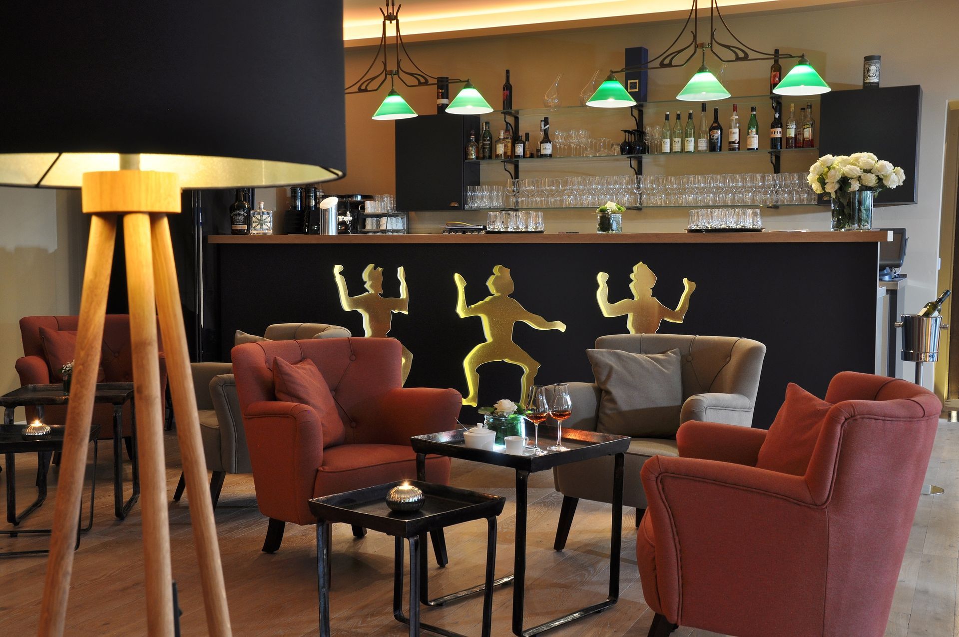 A cozy hotel bar with orange armchairs, small tables, a wooden floor, and decorative figurines on the dark counter.