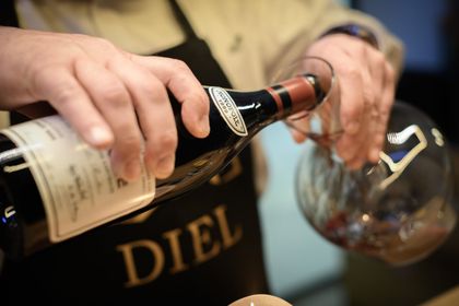 Eine Person, die eine schwarze Schürze mit der Aufschrift „DIEL“ trägt, gießt Rotwein aus einer Glasflasche in eine Karaffe aus klarem Glas.