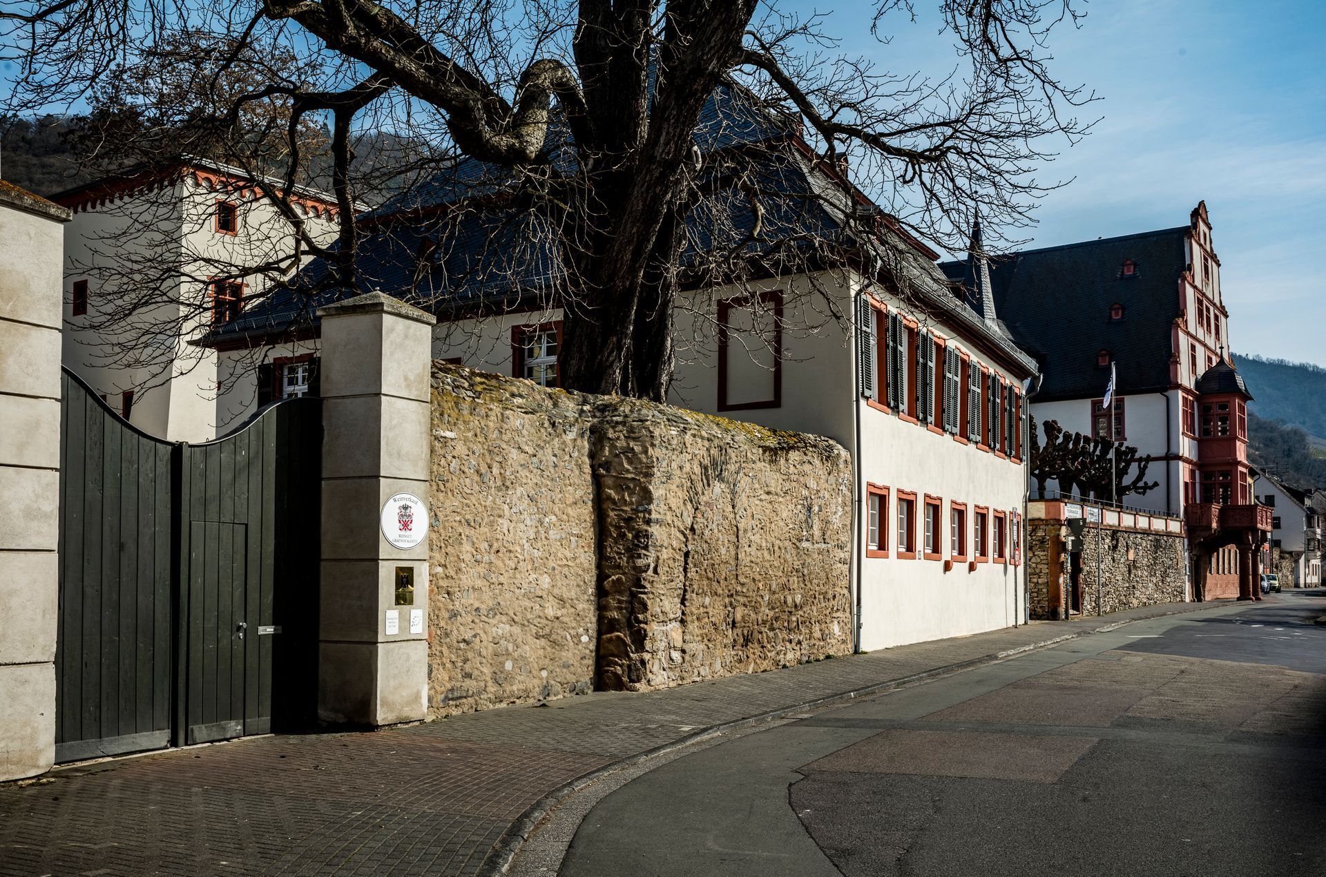 Ein historisches Steingebäude mit dunklem Tor und Baum an einer sonnigen, gepflasterten Straße in einer kleinen europäischen Stadt.