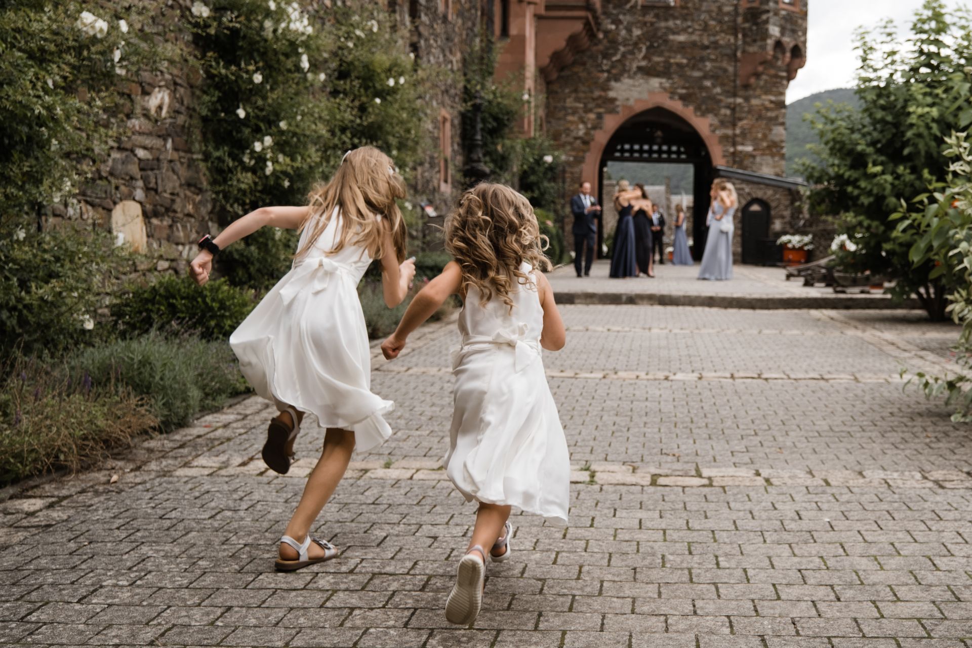 Zwei Kinder in weißen Kleidern rennen einen Steinpfad entlang auf einen Steinbogen zu, wo sich Gäste versammeln.