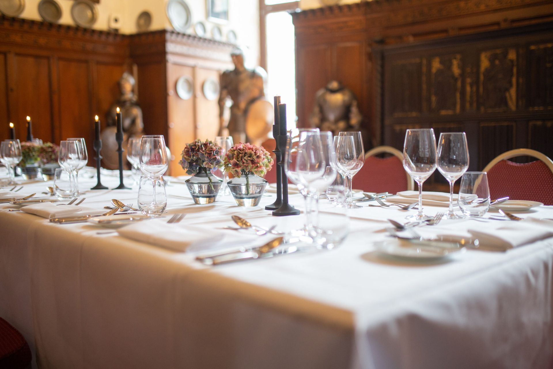 Ein langer, formeller Esstisch mit weißer Tischwäsche, Gläsern, Blumengestecken und Kerzen in einem rustikalen, holzgetäfelten Raum.