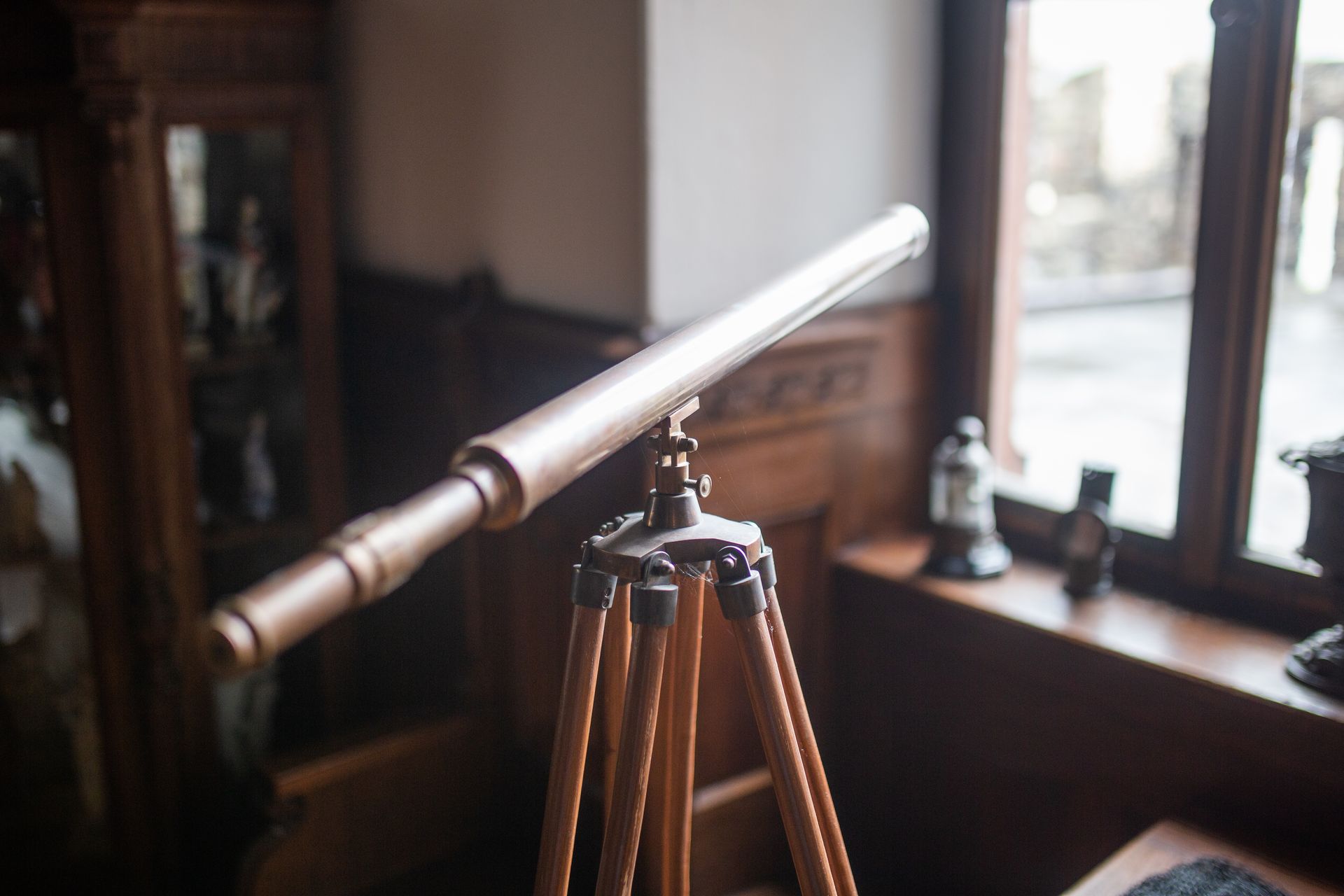 In a wood-paneled room with a window, there is an antique wooden telescope mounted on a tripod.