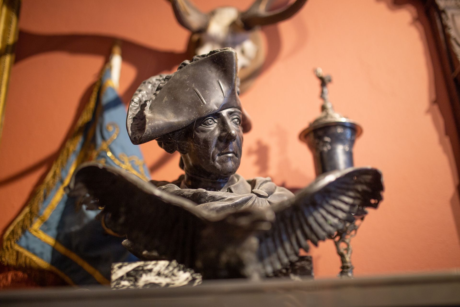 A bronze bust of a man wearing a tricorn hat, perched on an eagle with outstretched wings, set against a reddish wall, with a flag beside it.