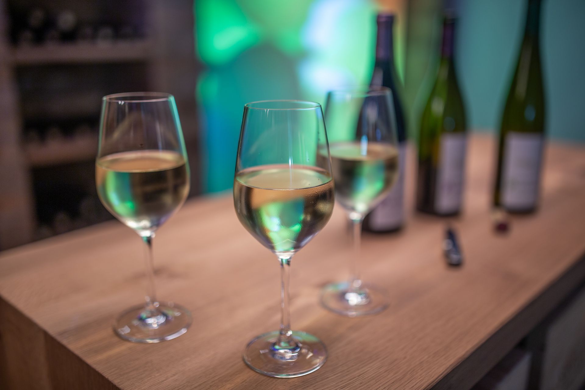 Drei mit Weißwein gefüllte Weingläser auf einem Holztisch, im verschwommenen Hintergrund Weinflaschen.