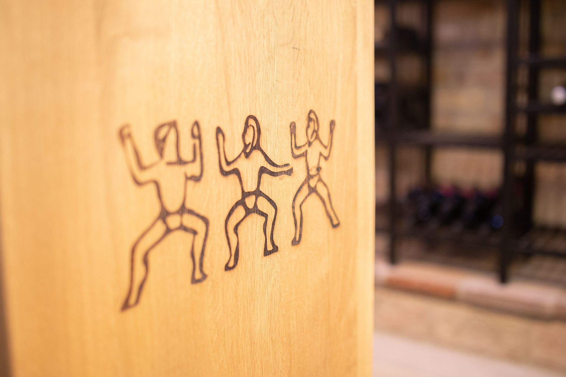 Nahaufnahme einer Holzoberfläche mit einer dunklen Strichzeichnung von drei stilisierten menschlichen Figuren in einer tänzerischen Pose.