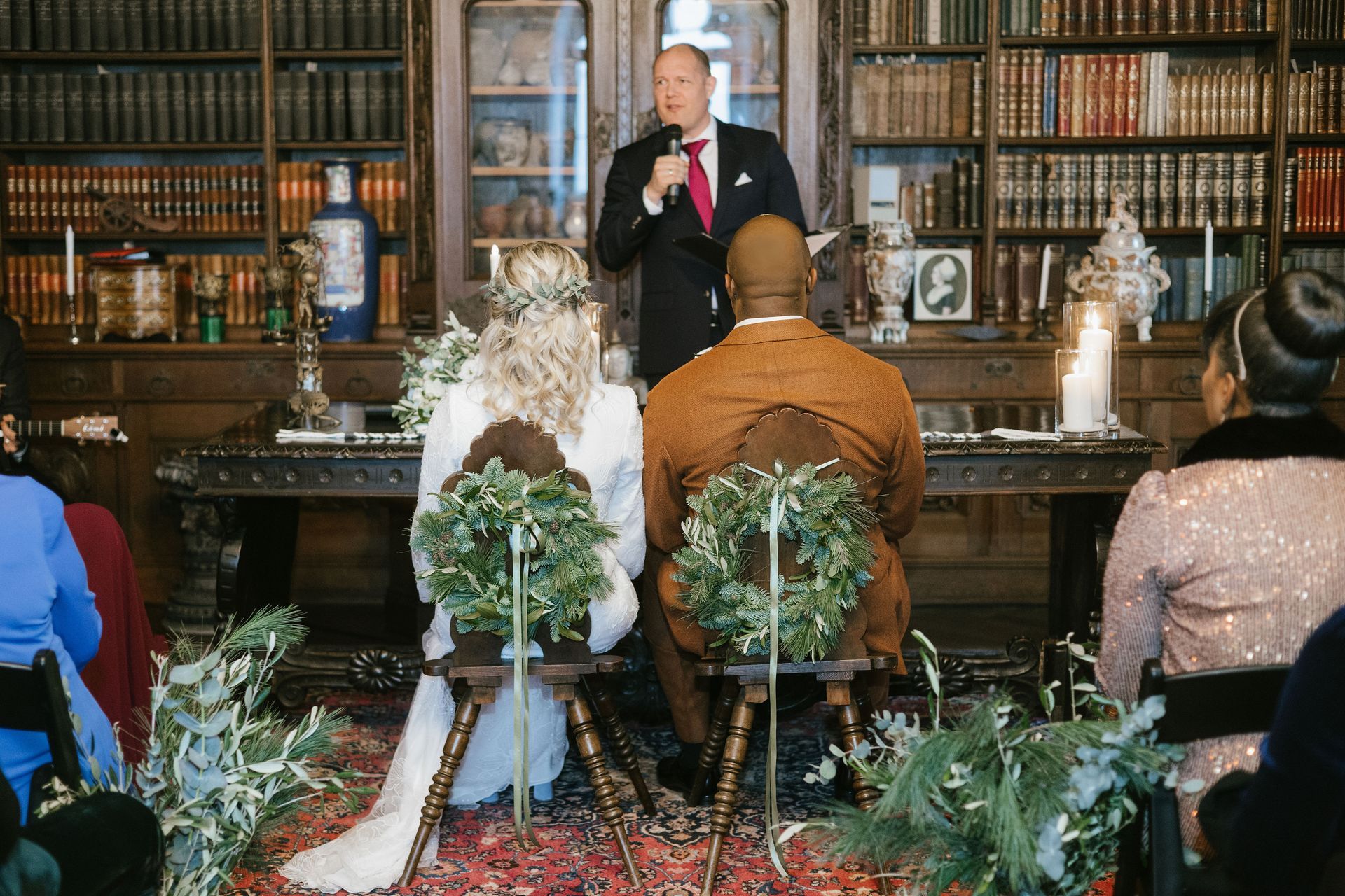 A wedding ceremony is taking place in a library, where a speaker is addressing a couple seated on chairs decorated with wreaths.