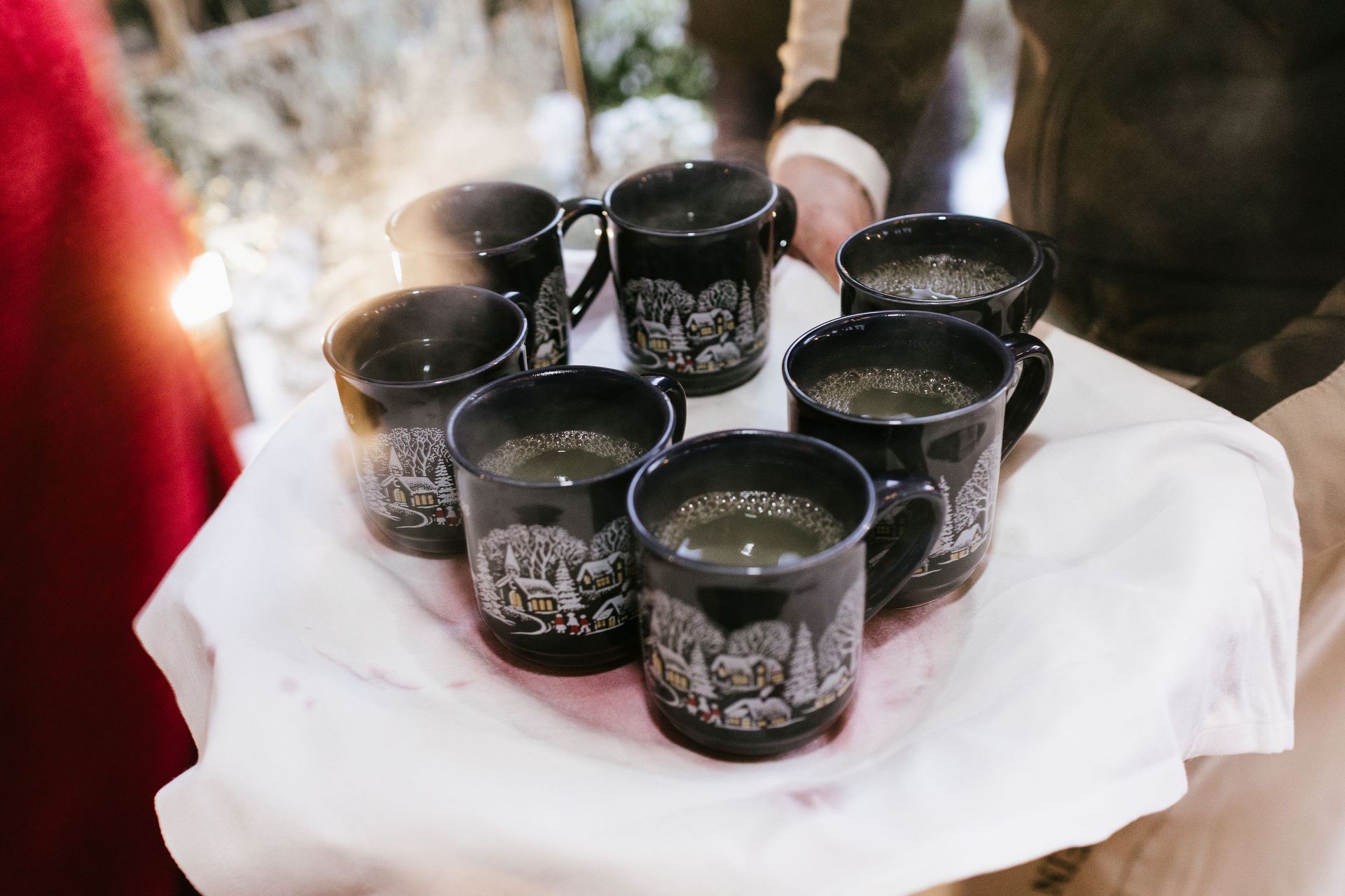 Eine Person hält ein mit einem weißen Tuch bedecktes Tablett, auf dem sieben schwarze Becher mit einem heißen, hellen Getränk stehen.