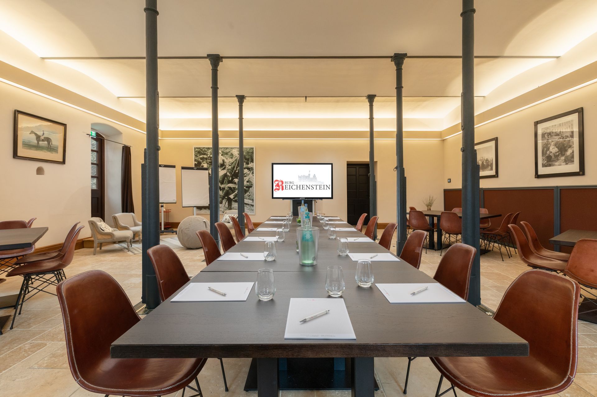 A formal meeting room with a long wooden table, surrounded by leather armchairs, bathed in soft light, and featuring exposed columns.