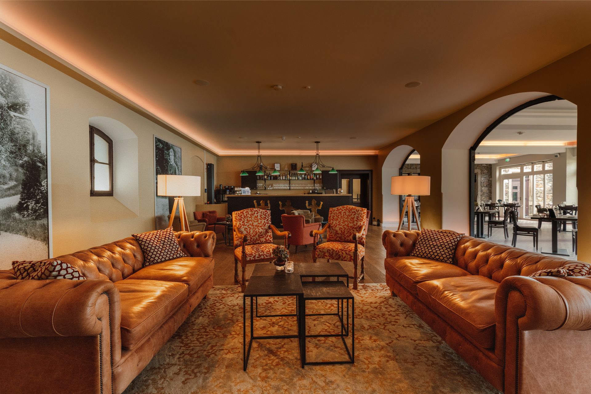 Eine Hotellobby mit zwei hellbraunen Ledersofas, gemusterten Sesseln und einem zentralen Tisch, die zu einem Essbereich führt.