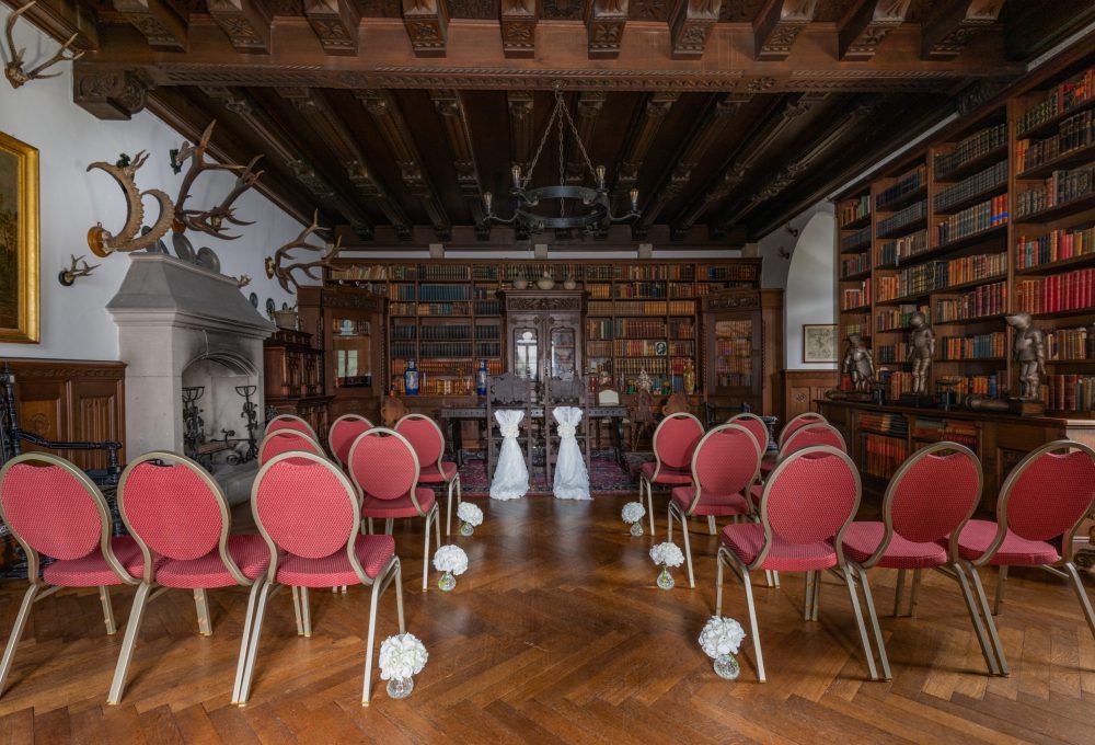 Ein für eine Hochzeitszeremonie hergerichteter Bibliotheksraum mit Reihen roter Stühle, denen zwei mit weißem Stoff bedeckte Stühle gegenüberstehen.