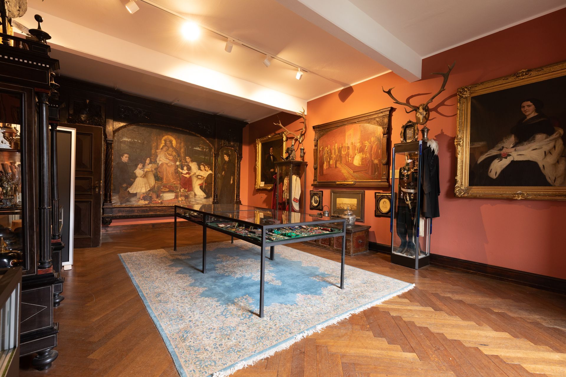 A room in a historical museum featuring paintings, a display case on a carpet, hardwood floors, and warm-toned walls.