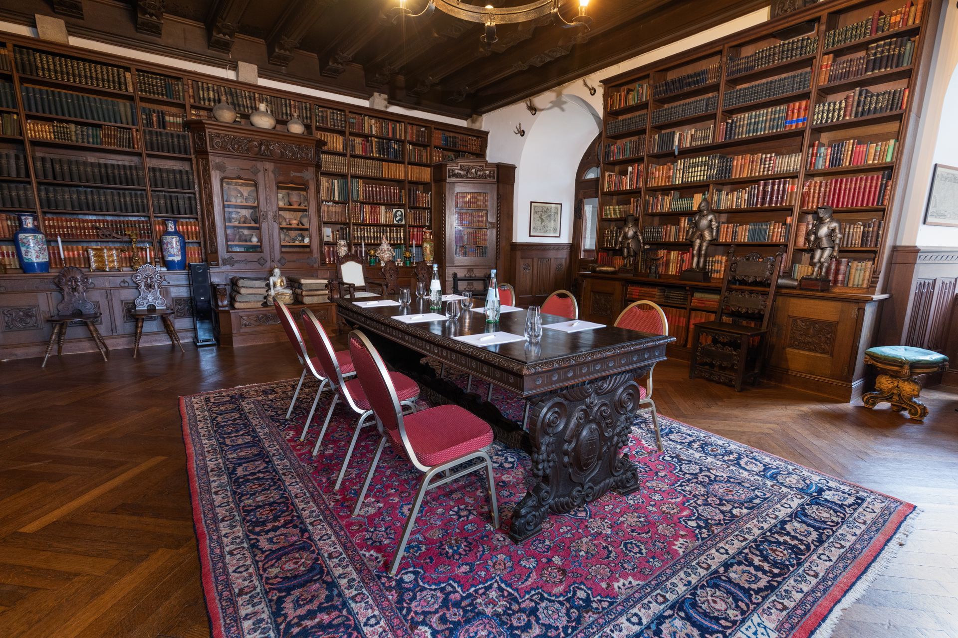 In a library room with dark wooden shelves and a beamed ceiling, there is a long wooden table and chairs on a patterned rug.