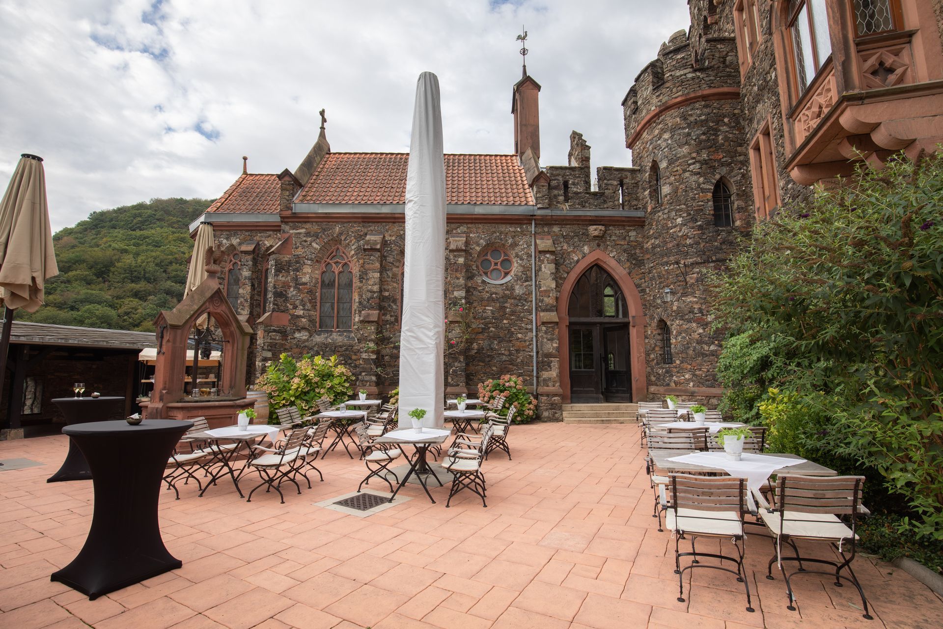 A terrace with tables and chairs in front of a stone building featuring a castle-like tower and a terracotta roof.