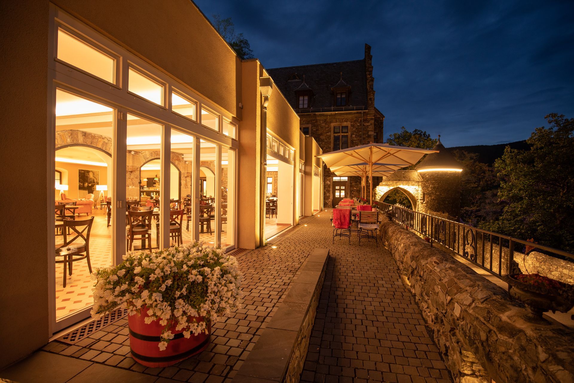 Terrasse im Freien bei Abenddämmerung mit Kopfsteinpflasterweg, Pflanzkasten und einem sanft beleuchteten Restaurantbereich.