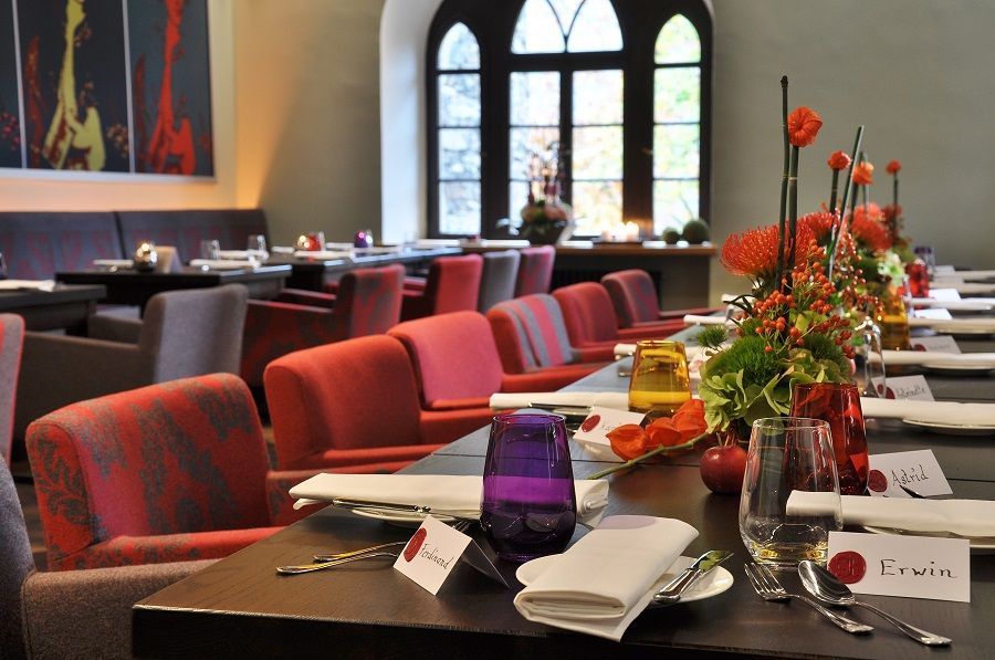In a restaurant, a long dining table has been set for an event, complete with place cards, colorful glasses, and floral arrangements.