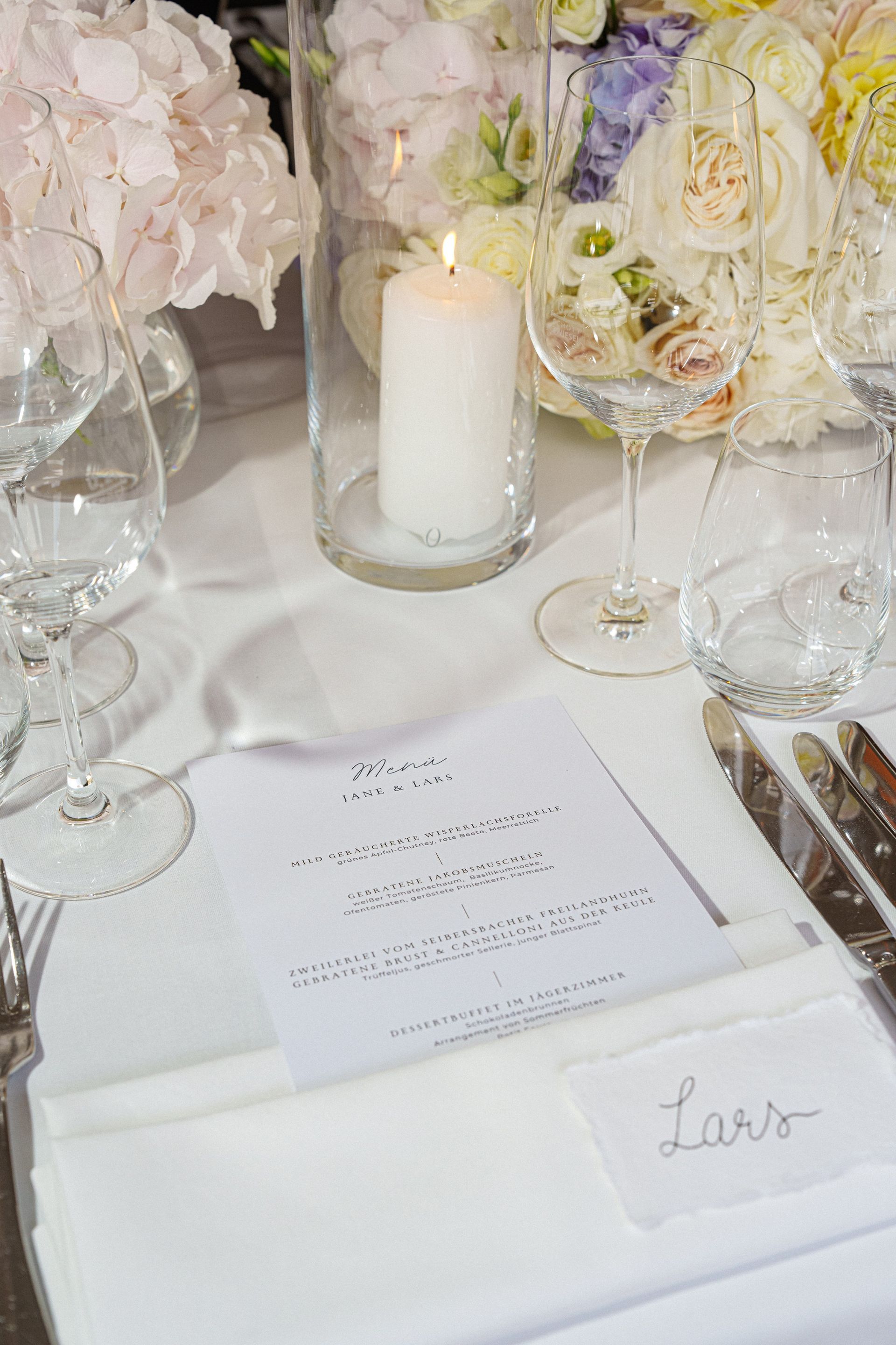 A place setting at a formal event featuring a printed menu, a candle, glasses, and a name card reading “Lars.”