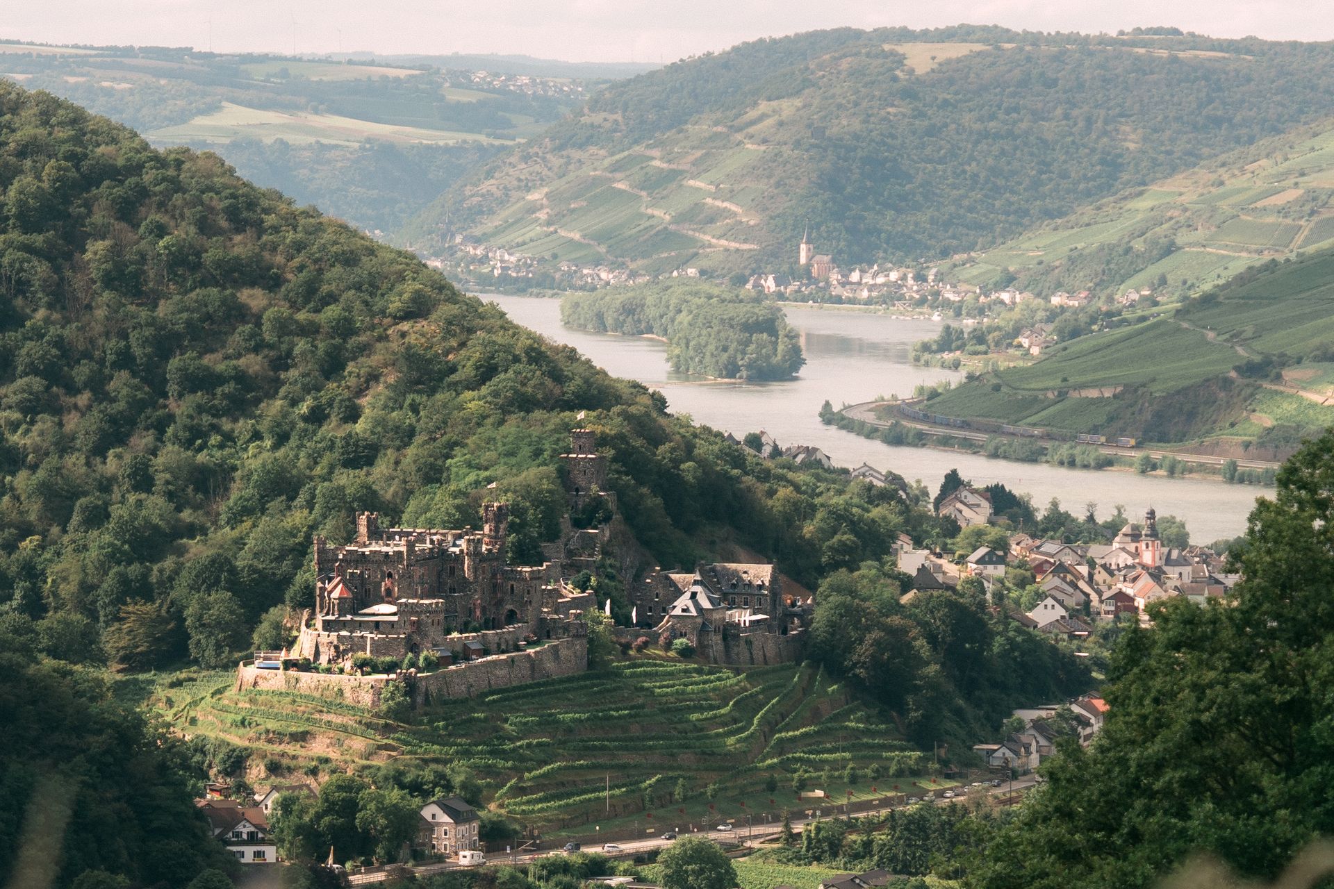 Auf einem bewaldeten Hügelhang thront eine steinerne Burg mit Blick auf einen gewundenen Fluss und ein kleines Dorf in einem Tal.