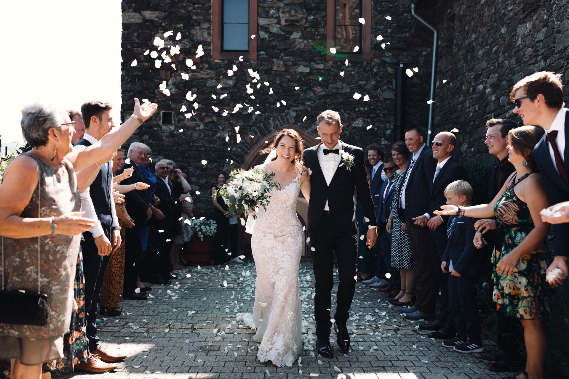 A bride and groom walk side by side in front of a stone building while guests throw confetti at them to celebrate.