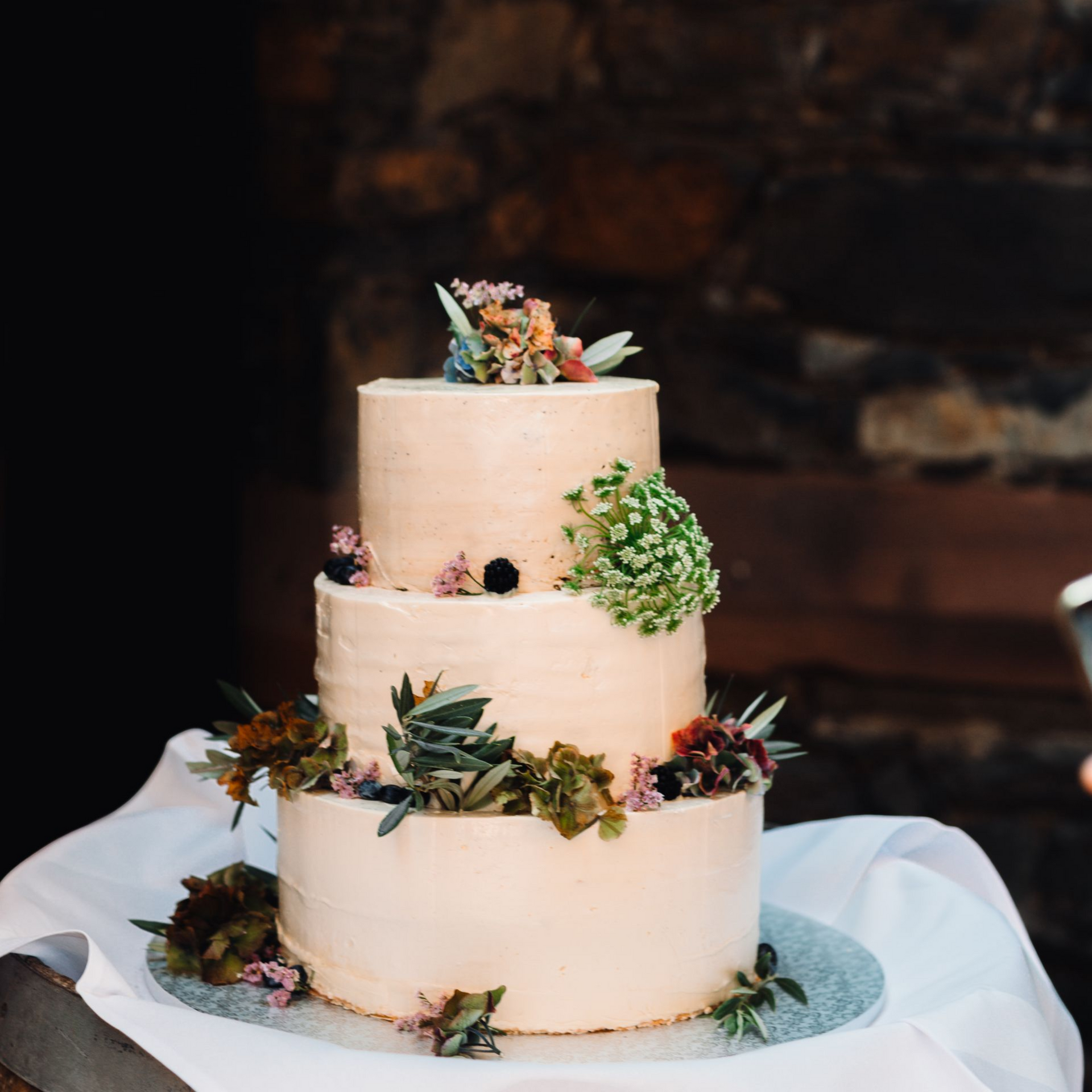 Eine dreistöckige, hellpfirsichfarbene Hochzeitstorte, verziert mit frischen Blumen, Grünzeug und Beeren, vor einer Steinmauer.