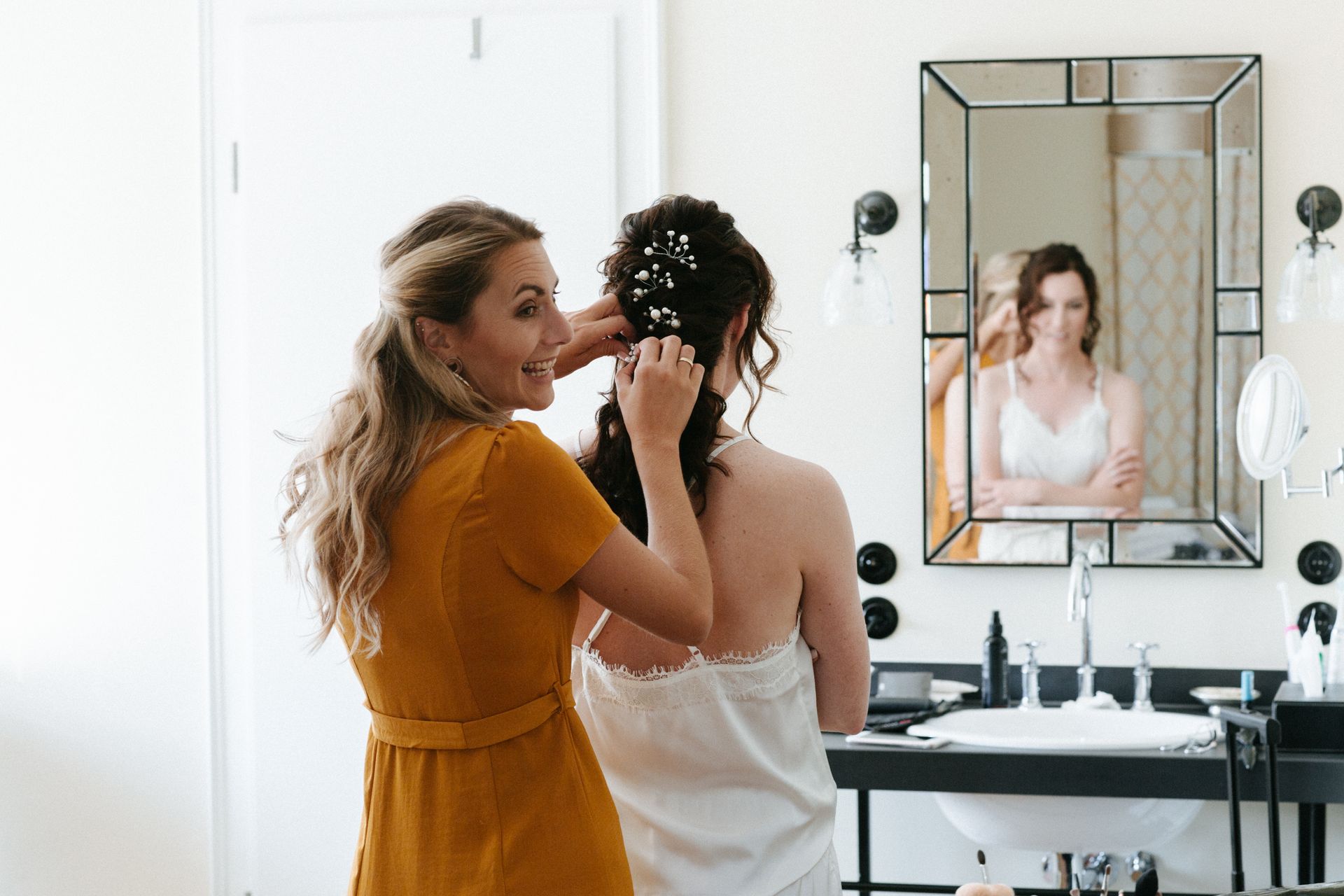 A person wearing a mustard-yellow dress is arranging small white flowers in the hair of another person wearing a white top, in front of a mirror.