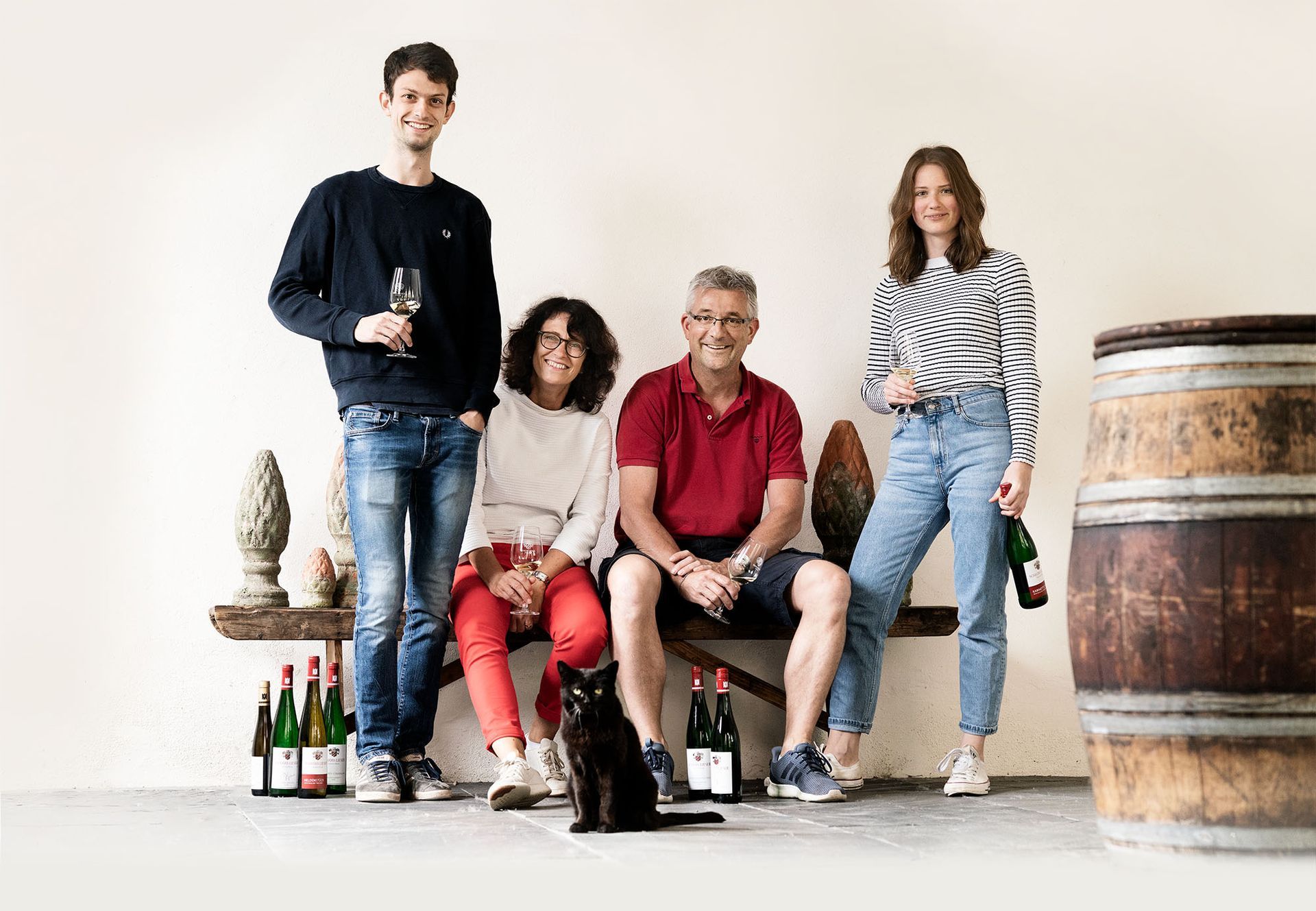 Vier Personen posieren in einem rustikalen Zimmer mit Weinflaschen, einem Fass und einer schwarzen Katze, die auf dem Boden sitzt.
