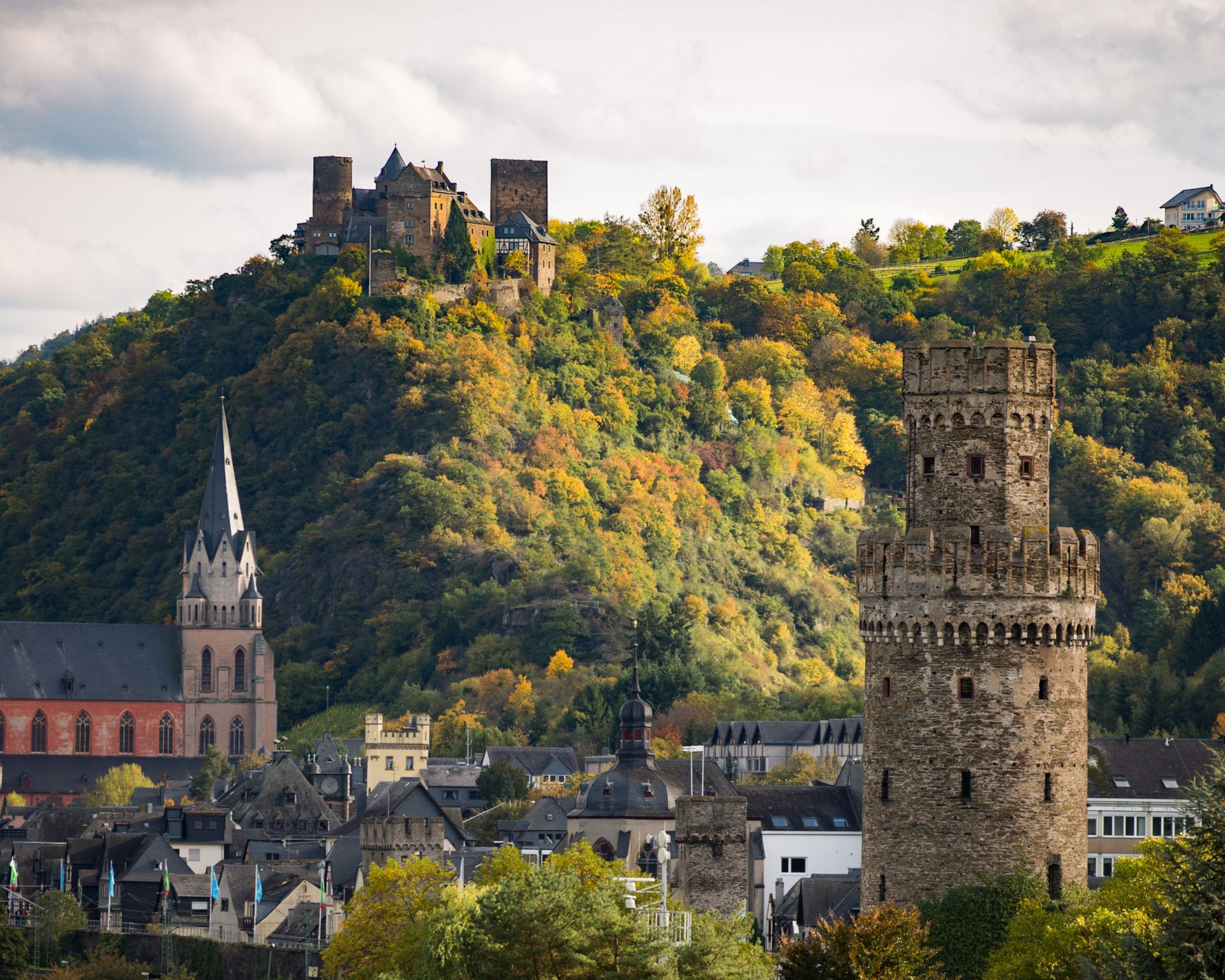 Im Herbst erhebt sich ein mittelalterlicher Steinturm vor einer Stadt mit einer gotischen Kirche und einer Burgruine auf einem bewaldeten Hügel.