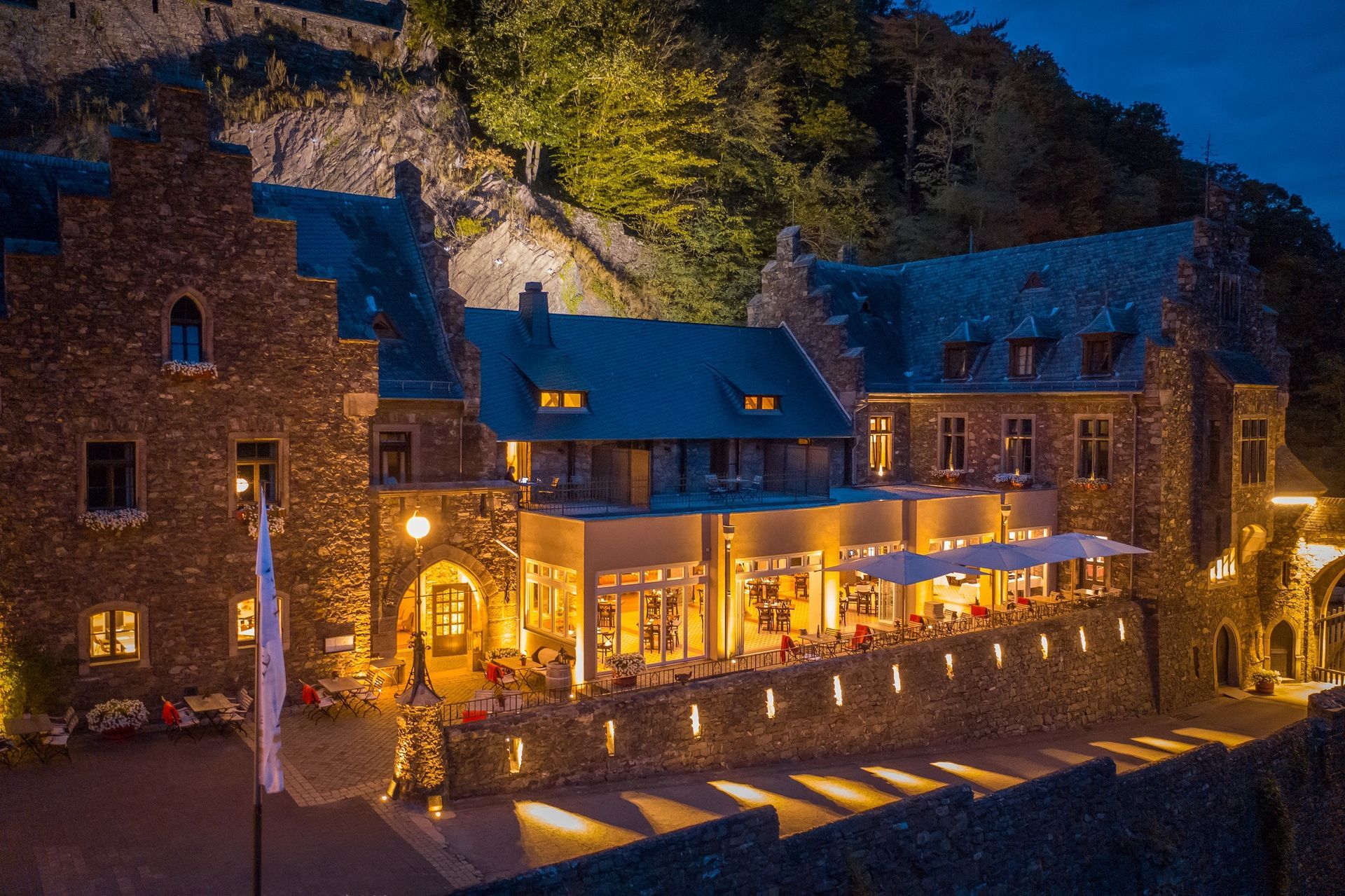 Ein aus Stein erbautes Schlosshotel, dessen Terrasse und Fassade in der Abenddämmerung vor einem dunklen Hang in warmes, goldenes Licht getaucht sind.