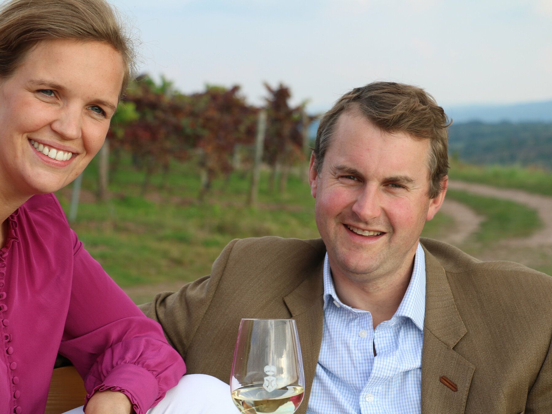 Ein lächelndes Paar posiert im Freien vor einem Weinberg; im Vordergrund ist ein Weinglas zu sehen.