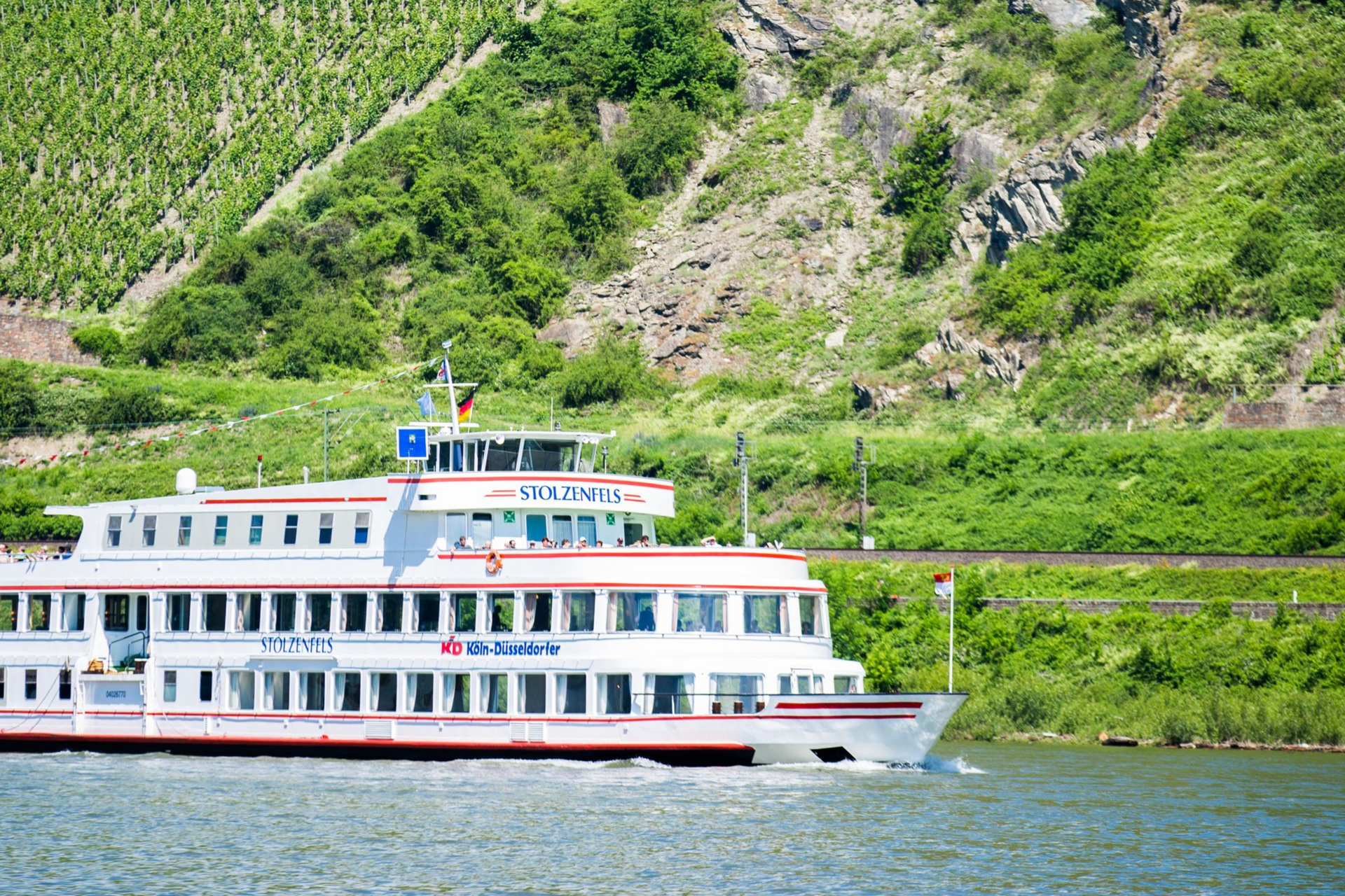 Ein weißes Kreuzfahrtschiff fährt auf einem Fluss vorbei an einem üppig bewachsenen, terrassenförmig angelegten grünen Hügel in Deutschland.