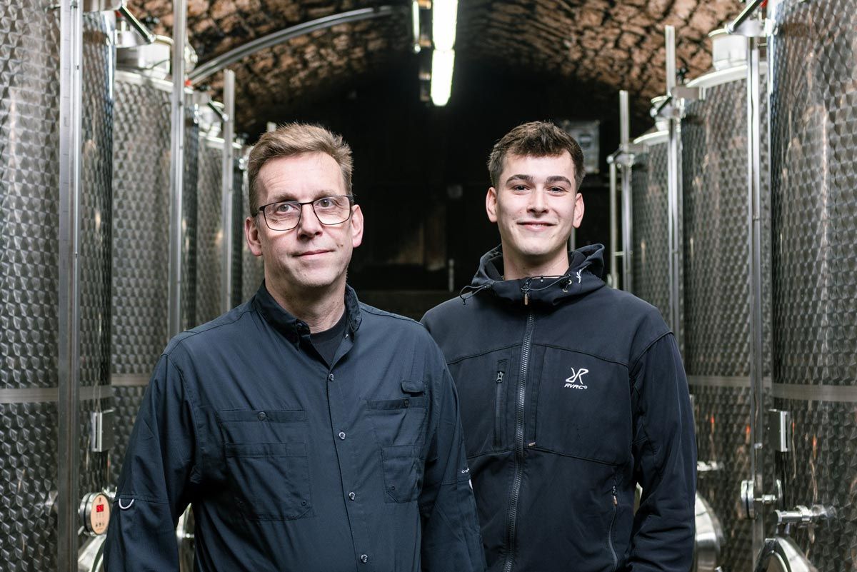 Zwei Personen stehen in einem Keller mit Steindecke, flankiert von Reihen hoher industrieller Metalltanks.
