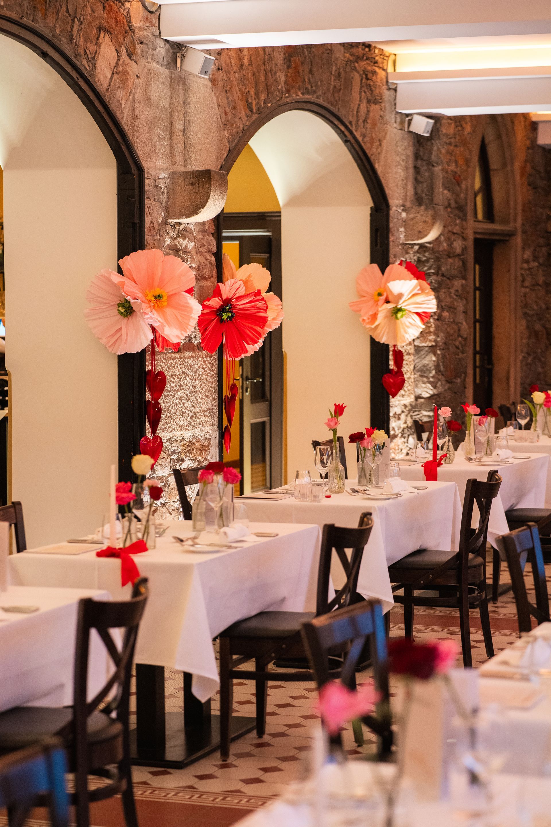 Tische mit weißen Tischdecken und großen Papierblumen-Gestecken in einem gewölbten, von Steinmauern umgebenen Restaurantinterieur.