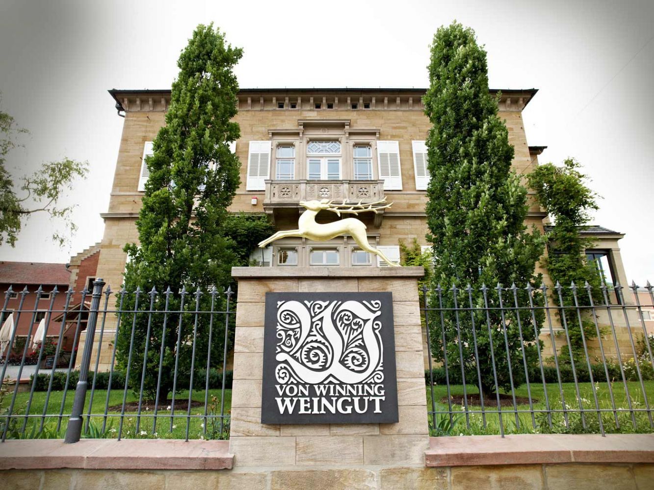 Ein Steinschild mit der Aufschrift „Von Winning Weingut“ vor einem imposanten Gebäude, flankiert von zwei hohen, schlanken immergrünen Bäumen.