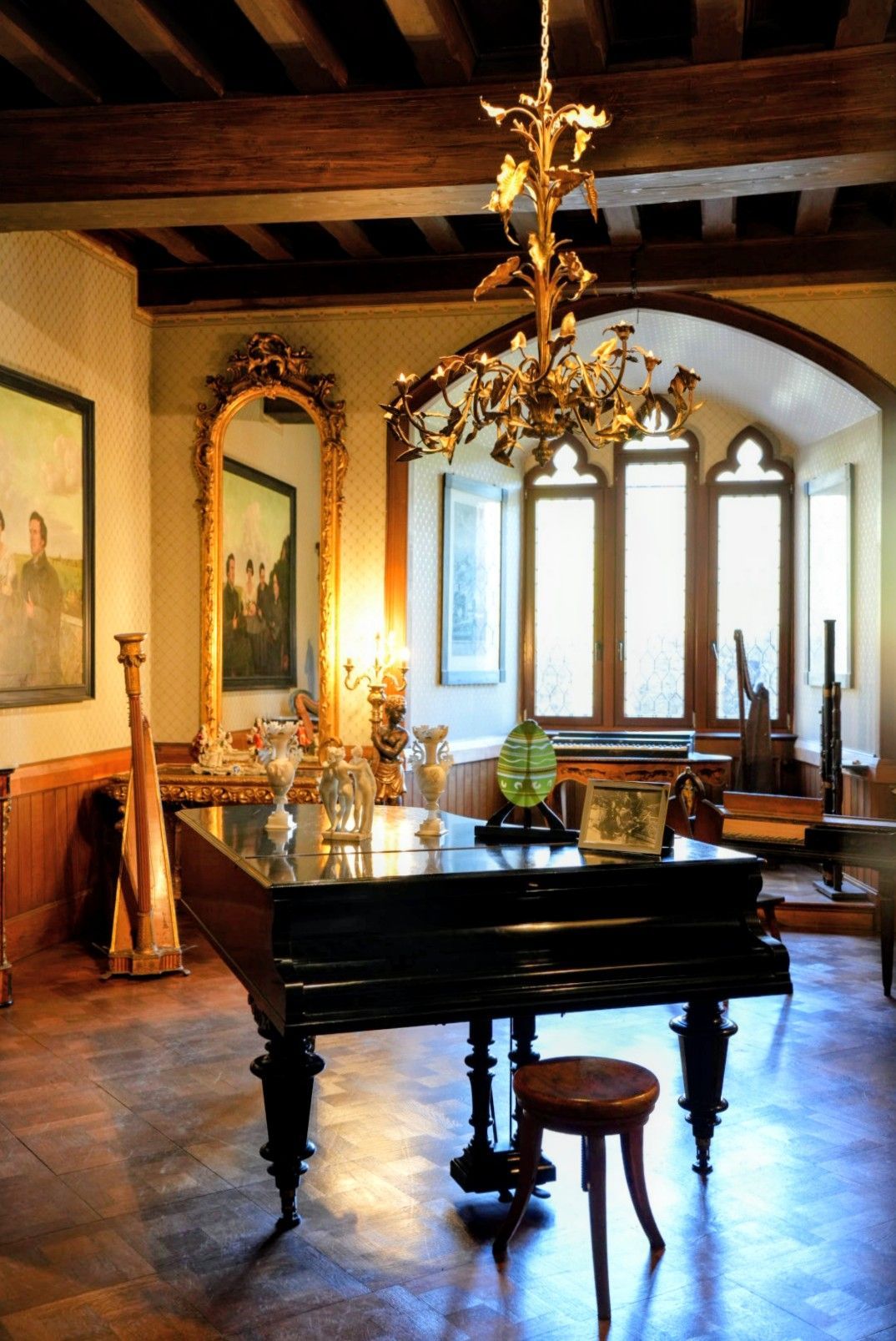 In einem Raum mit einem großen Kronleuchter, einem hohen Bogenspiegel, einer Harfe und dunklen Holzbalkendecken steht ein Flügel.