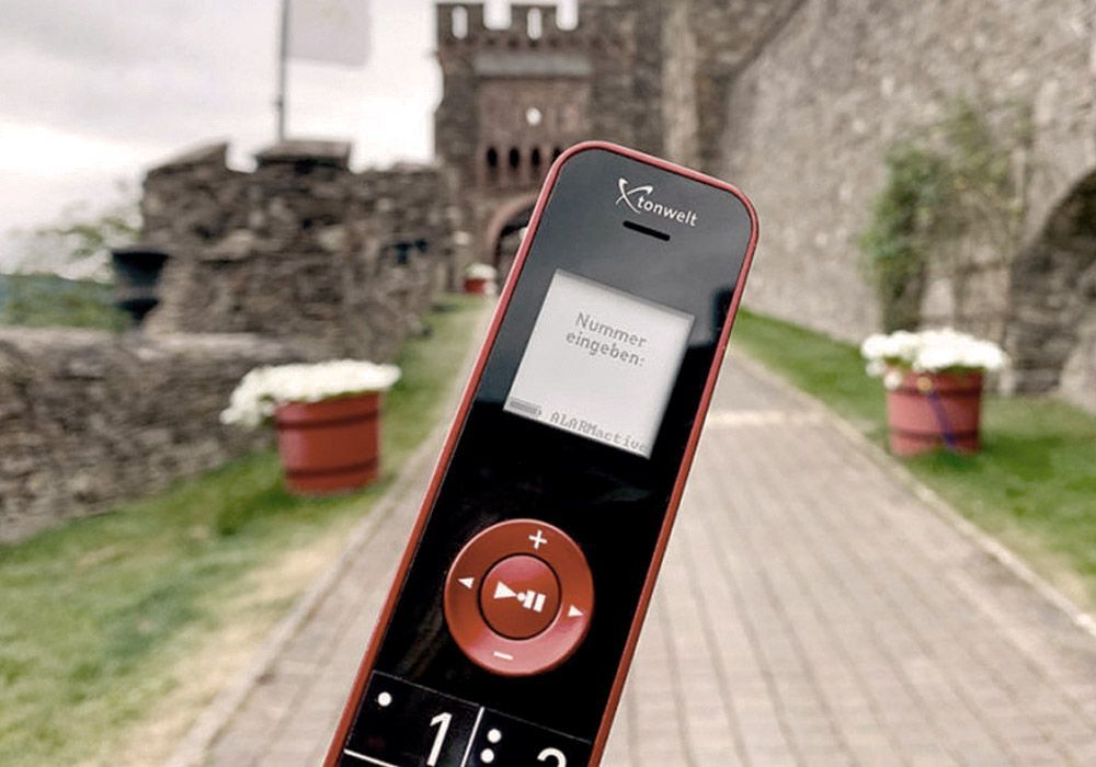 A portable audio guide device displays text in front of a stone castle gatehouse; along the path are brick planters.