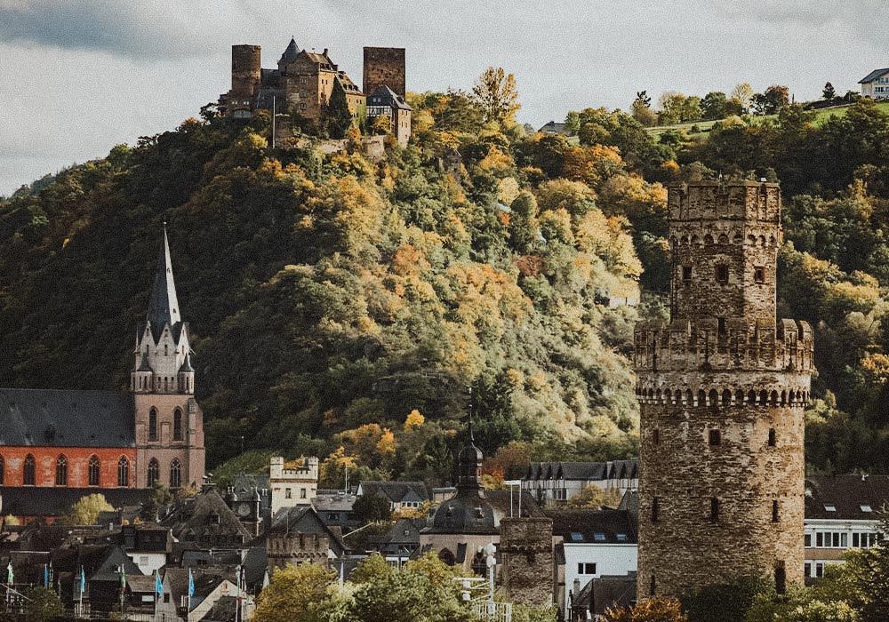 Eine mittelalterliche Steinburg überblickt ein malerisches Städtchen mit einer hohen Kirche und einem markanten Wachturm, der in einen bewaldeten Hügel eingebettet ist.