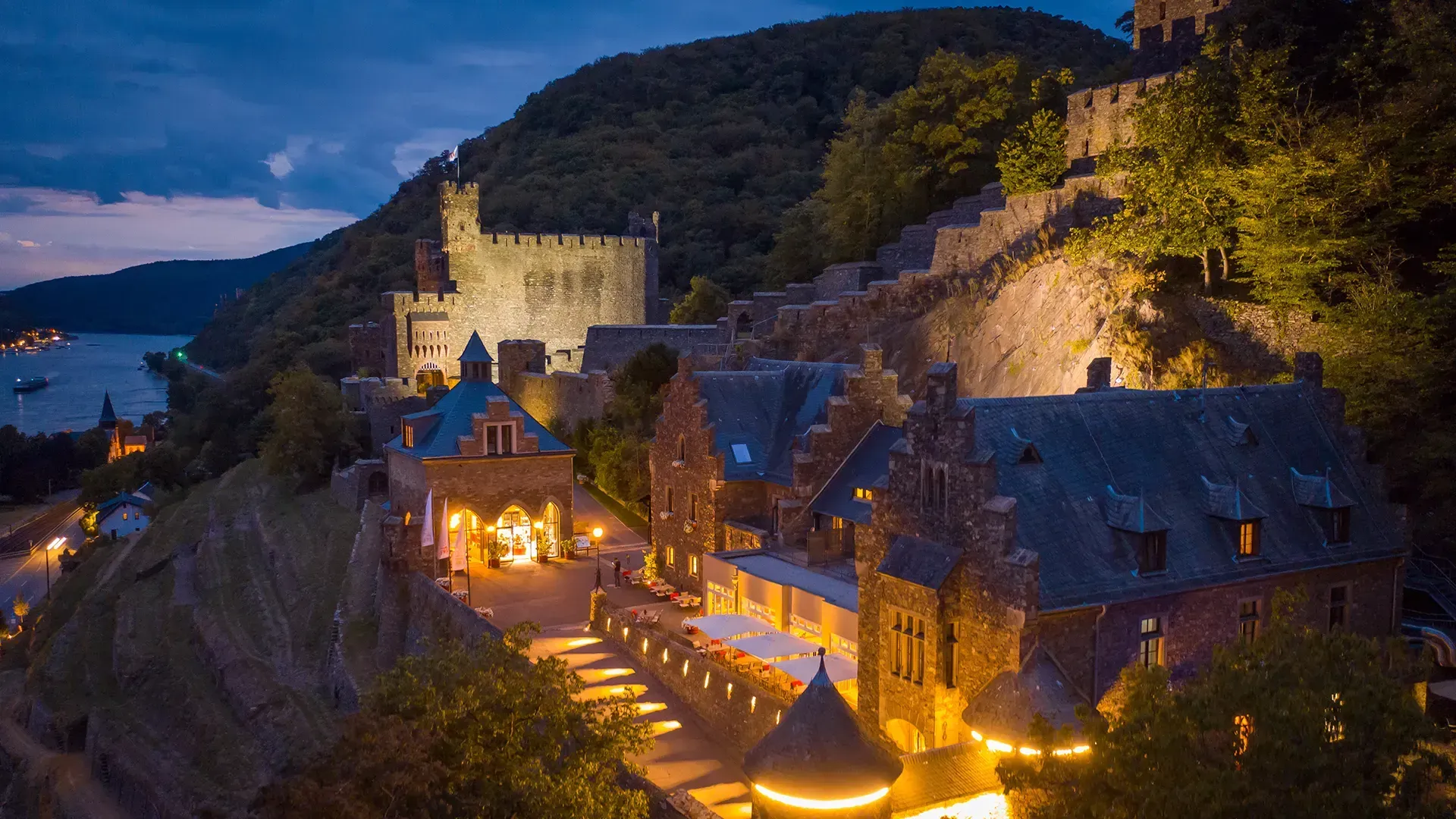 Eine beleuchtete historische Burg, die in der Abenddämmerung auf einer Felsklippe über einem Fluss thront.