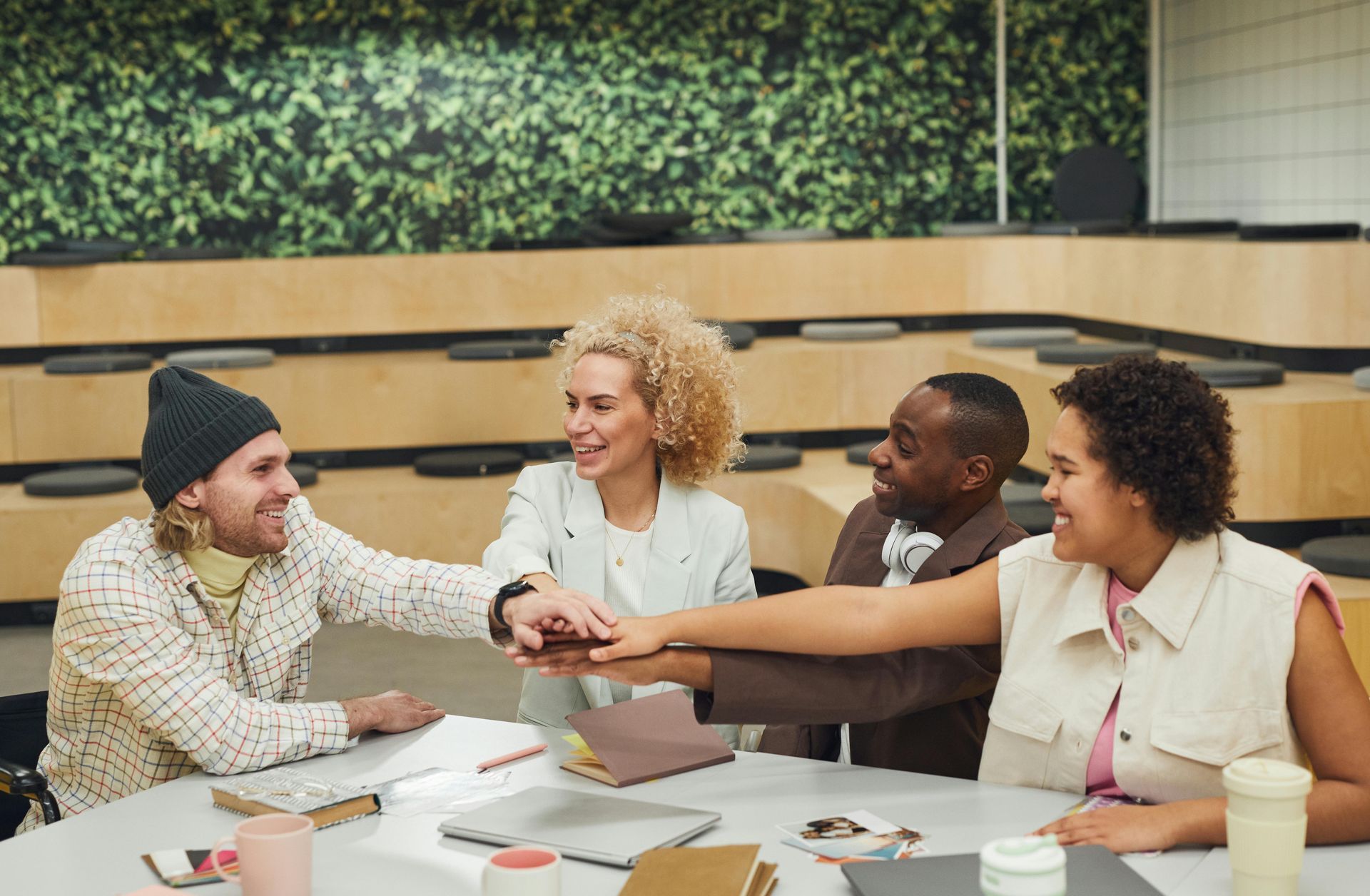 Vier Personen sitzen um einen Tisch in einem modernen Arbeitszimmer mit begrünten Wänden und lächeln, während sie sich an den Händen fassen.