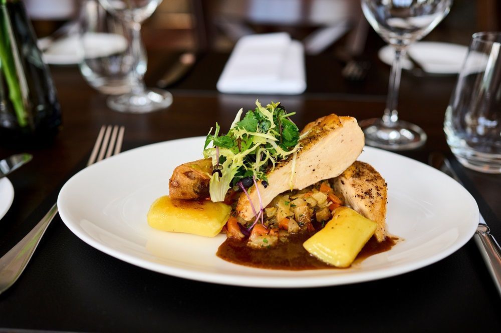 Ein Tellergericht aus Hühnerfleischscheiben, goldbraun frittierten Quadraten und Gemüse, serviert mit Soße auf einem dunklen Restauranttisch.
